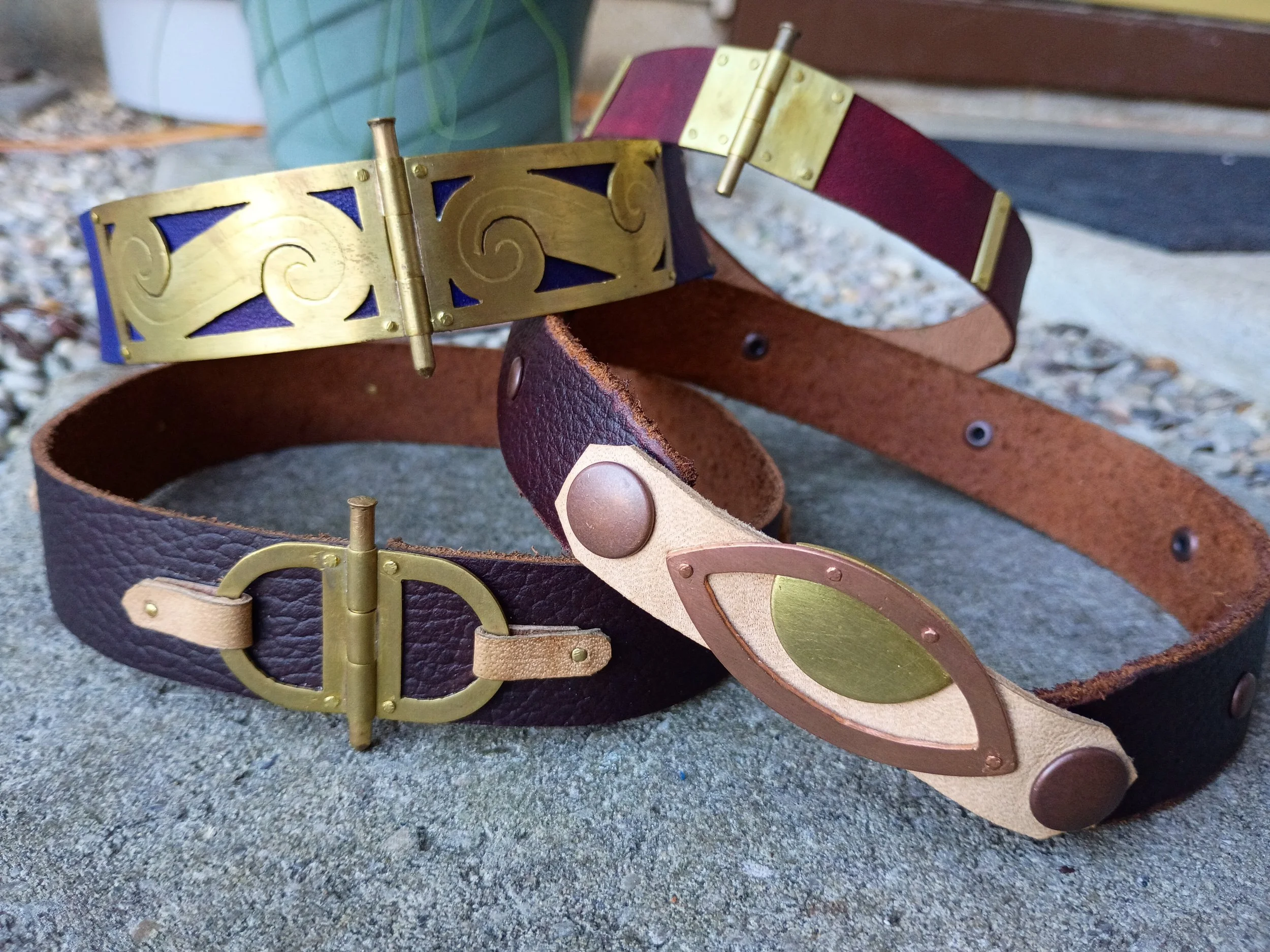 Two leather collars, one dark brown and the other tan, with decorative brass and metal clasps, lying on a concrete surface.