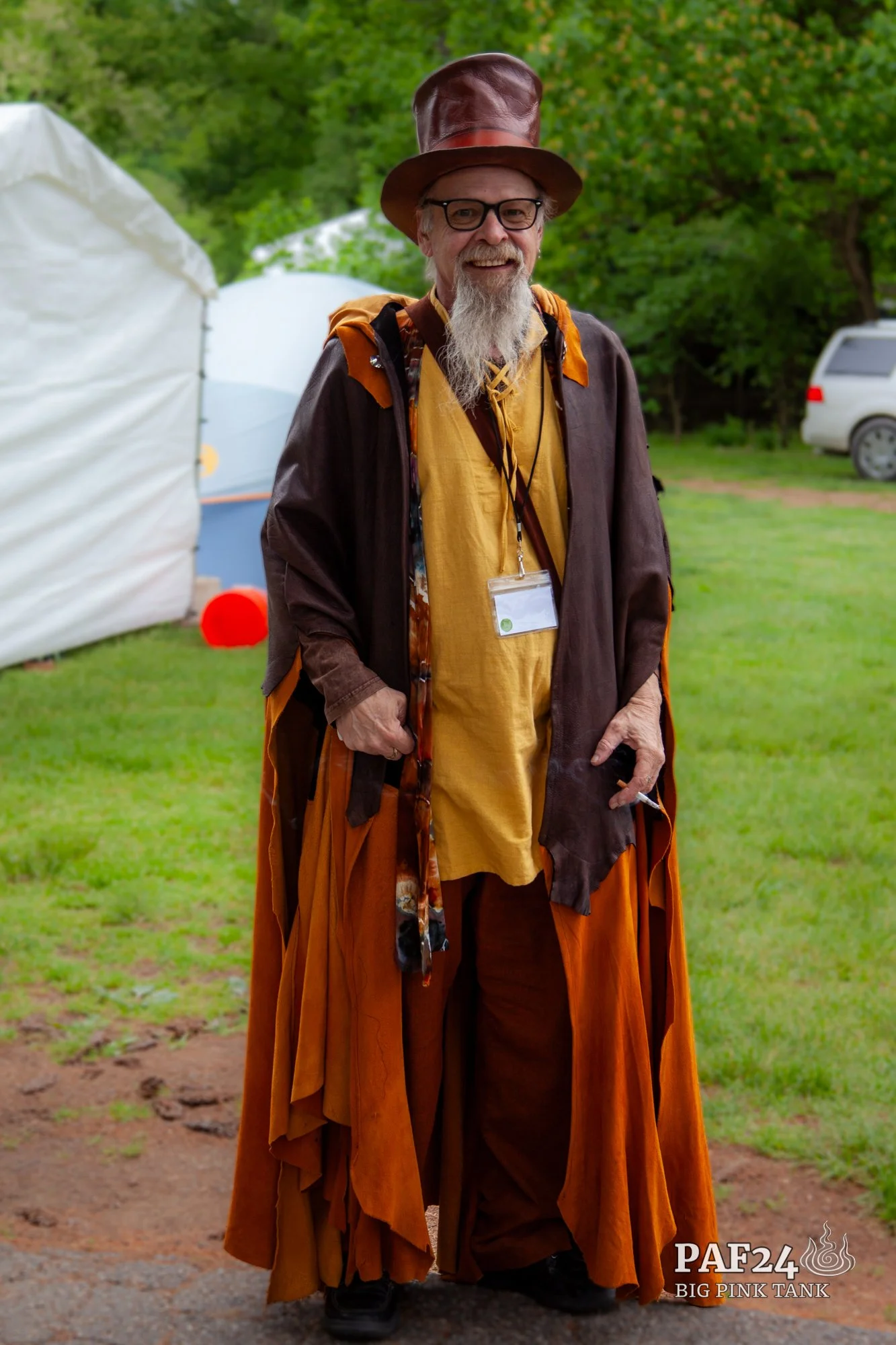 A man dressed in a colorful, layered costume inspired by a wizard or fantasy character, standing outdoors on grass with tents in the background.