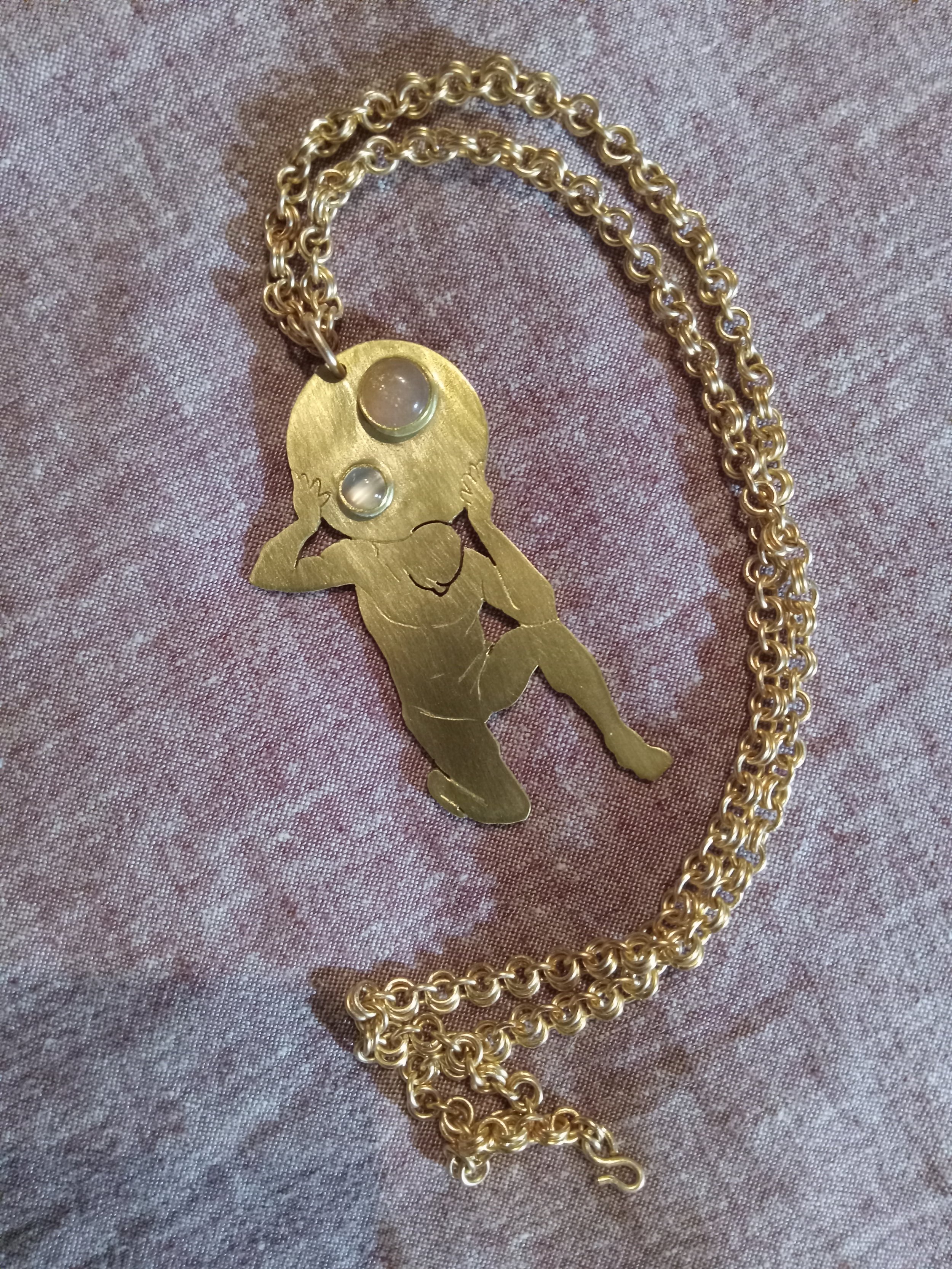 Gold necklace with a pendant depicting a stylized child with a round head, holding its head with both hands, placed on a textured pink fabric background.