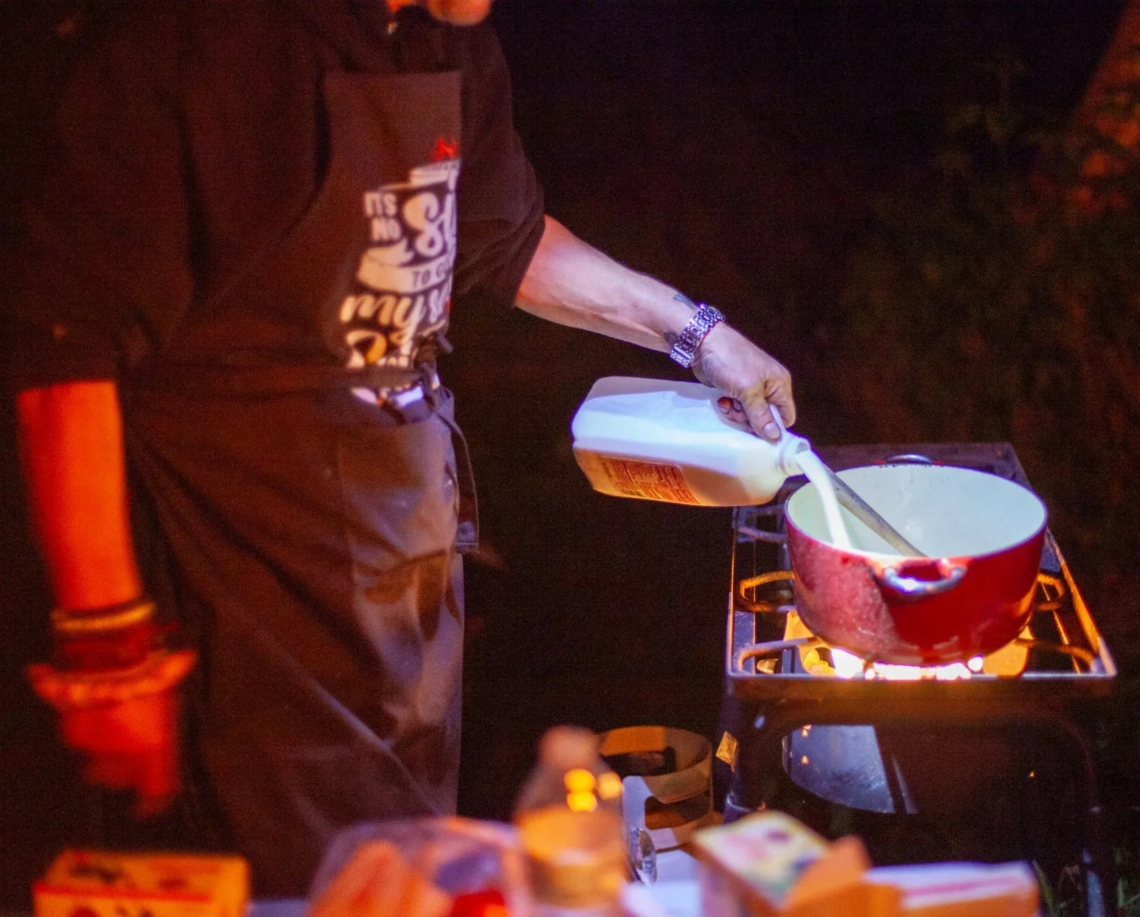 Person cooking on a stove at night, pouring milk into a red pot. The person is wearing a black shirt with writing on it, and a watch on their left wrist.