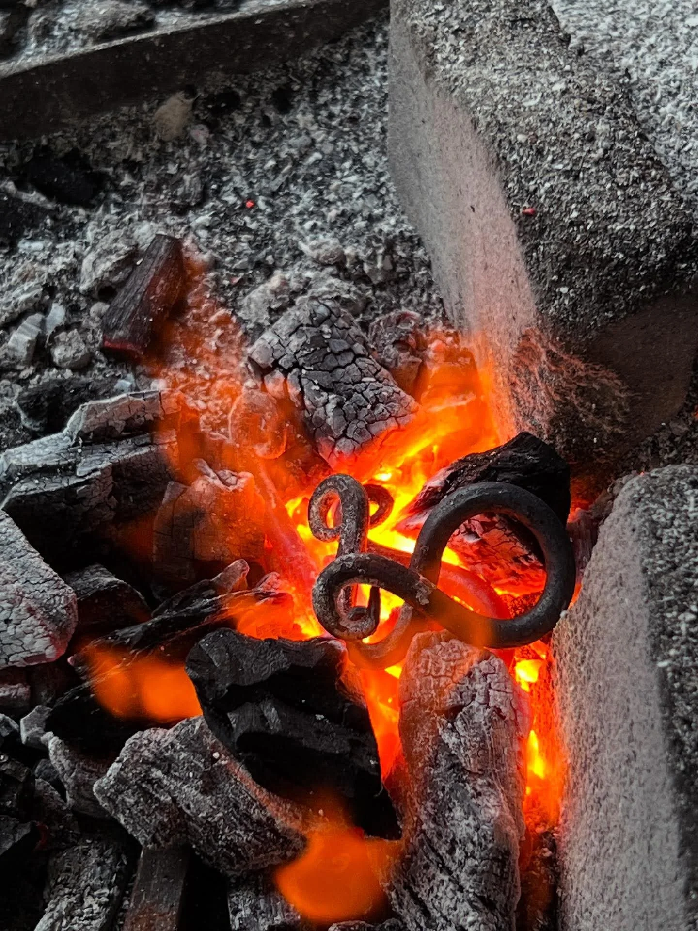 A close-up of glowing hot coals and burning wood in a fire pit with a piece of blackened wrought iron decorative piece among the embers.
