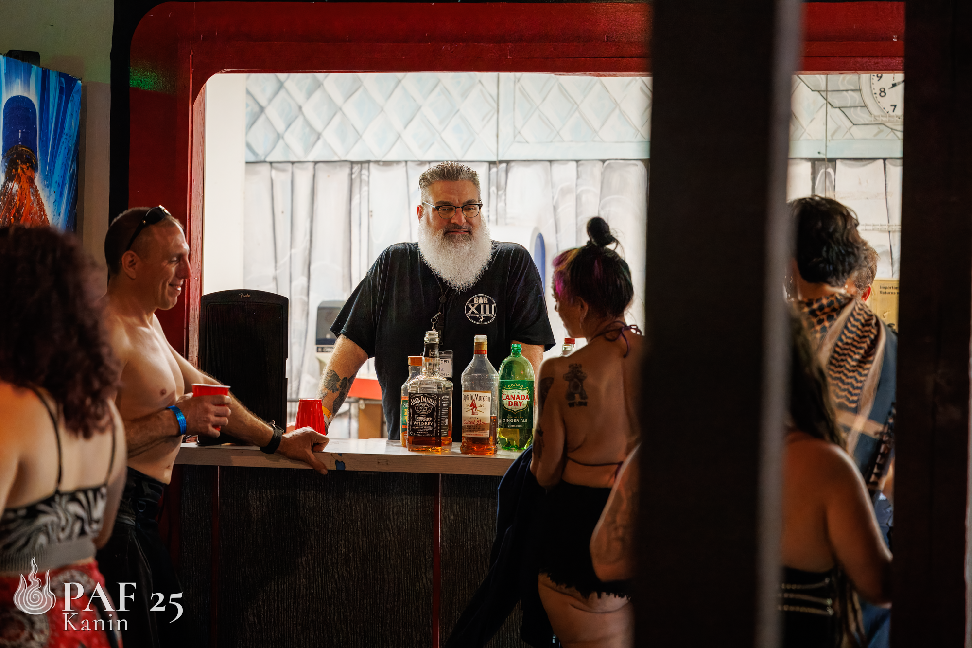 A bartender with a beard and glasses standing behind a bar counter, chatting with a woman with purple hair and tattoos. Several bottles of alcohol and red cups are on the bar. Other people are around, some shirtless and some with tattoos, in a lively social setting.