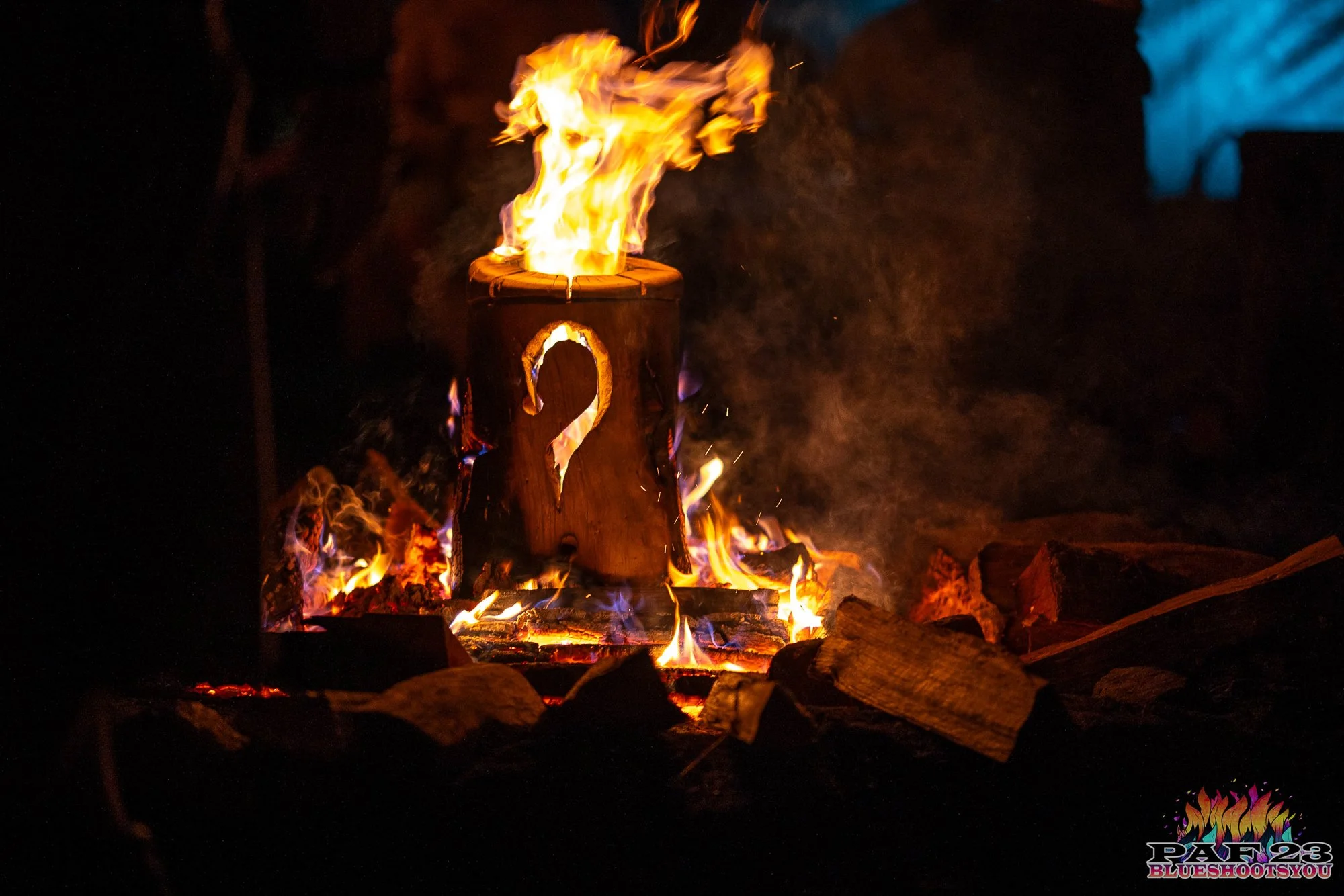 A wooden cauldron with a question mark cutout on its side is burning inside a campfire, with flames and embers around it at night.