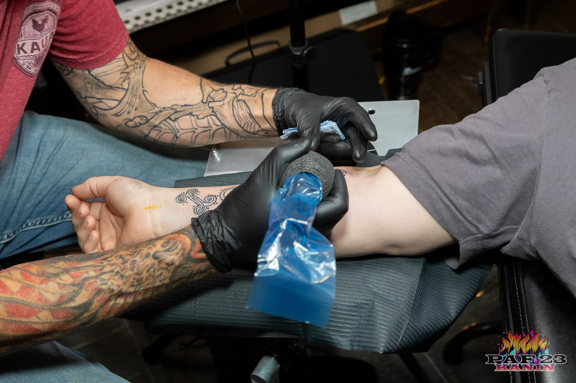 Tattoo artist in black gloves tattooing a person's forearm while seated on a black chair.