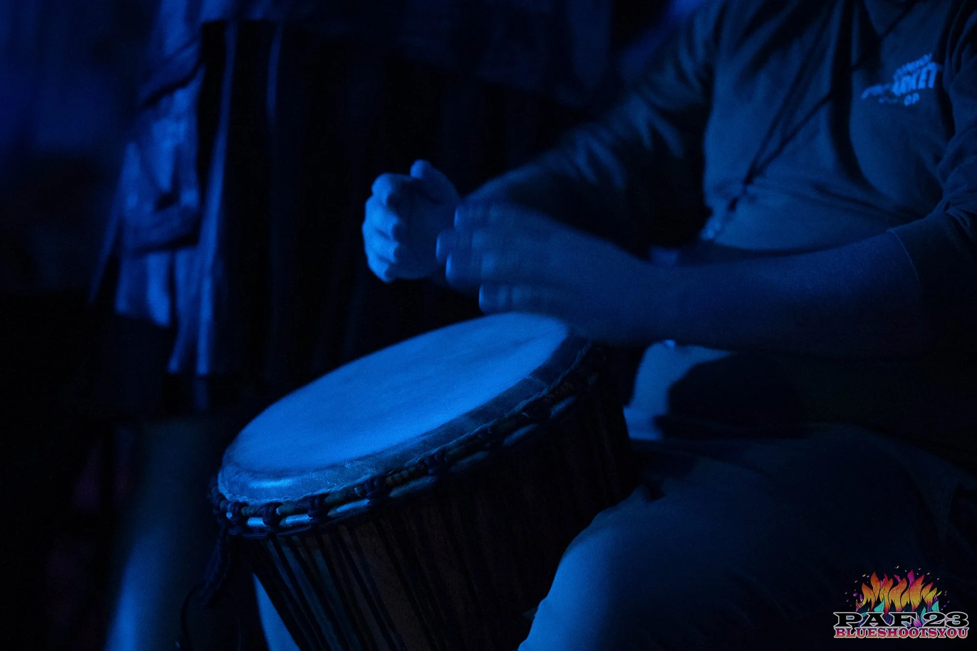 Person playing a hand drum, illuminated in blue lighting, wearing a long-sleeve shirt.