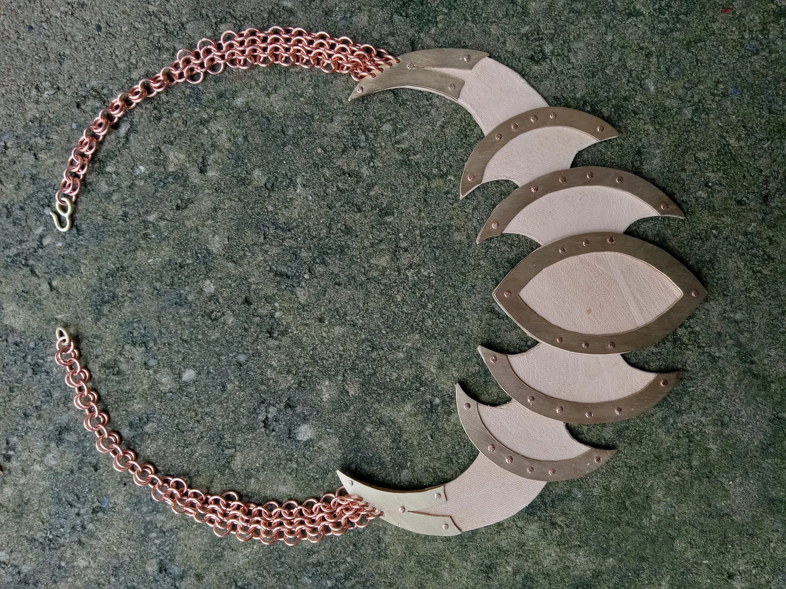 A necklace with a chain made of interlinked copper-colored rings and a pendant featuring a design of silver crescent shapes and beige leather accents arranged in a circular pattern.