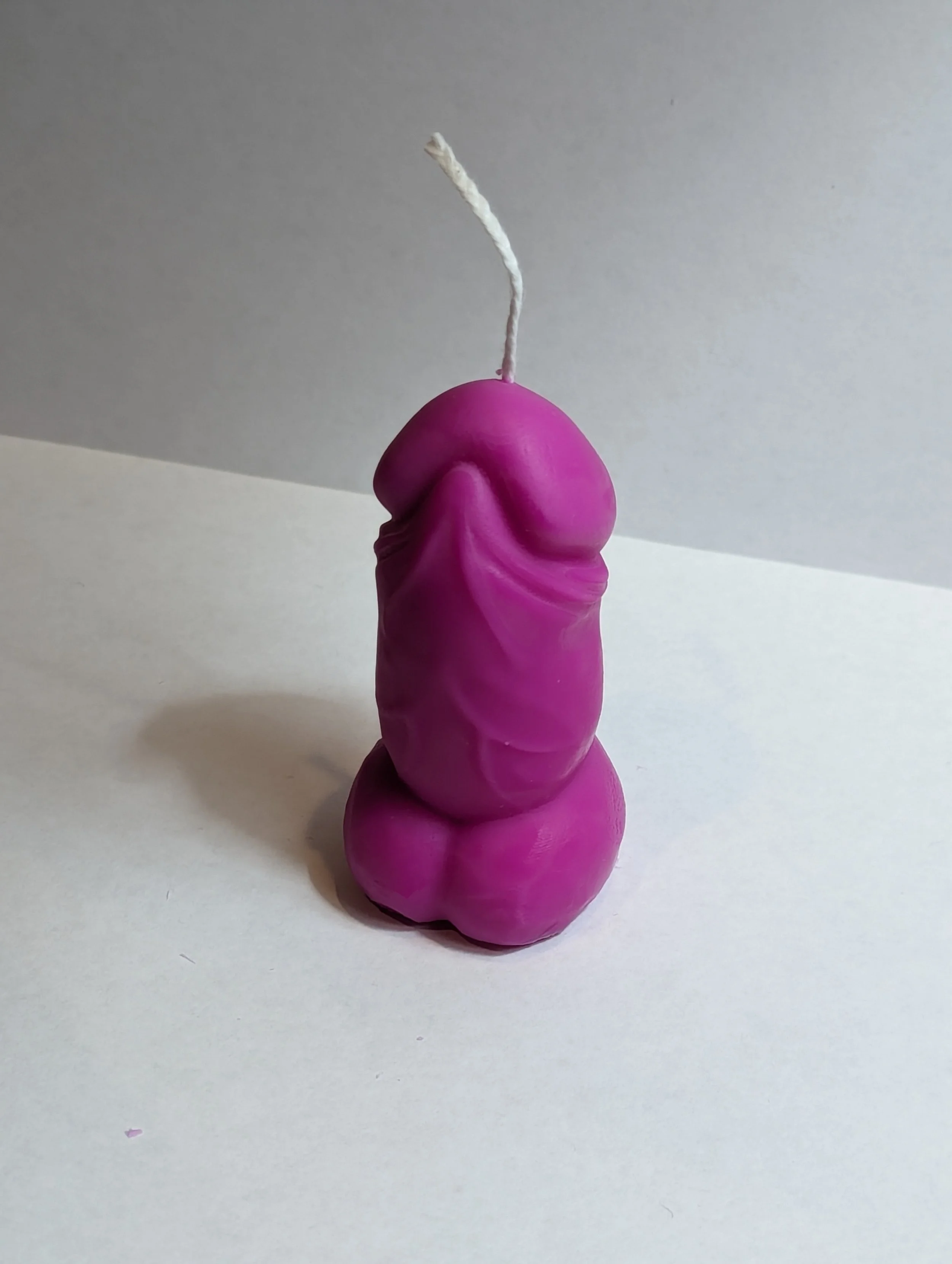 A purple wax candle in the shape of penis and a white wick standing upright.