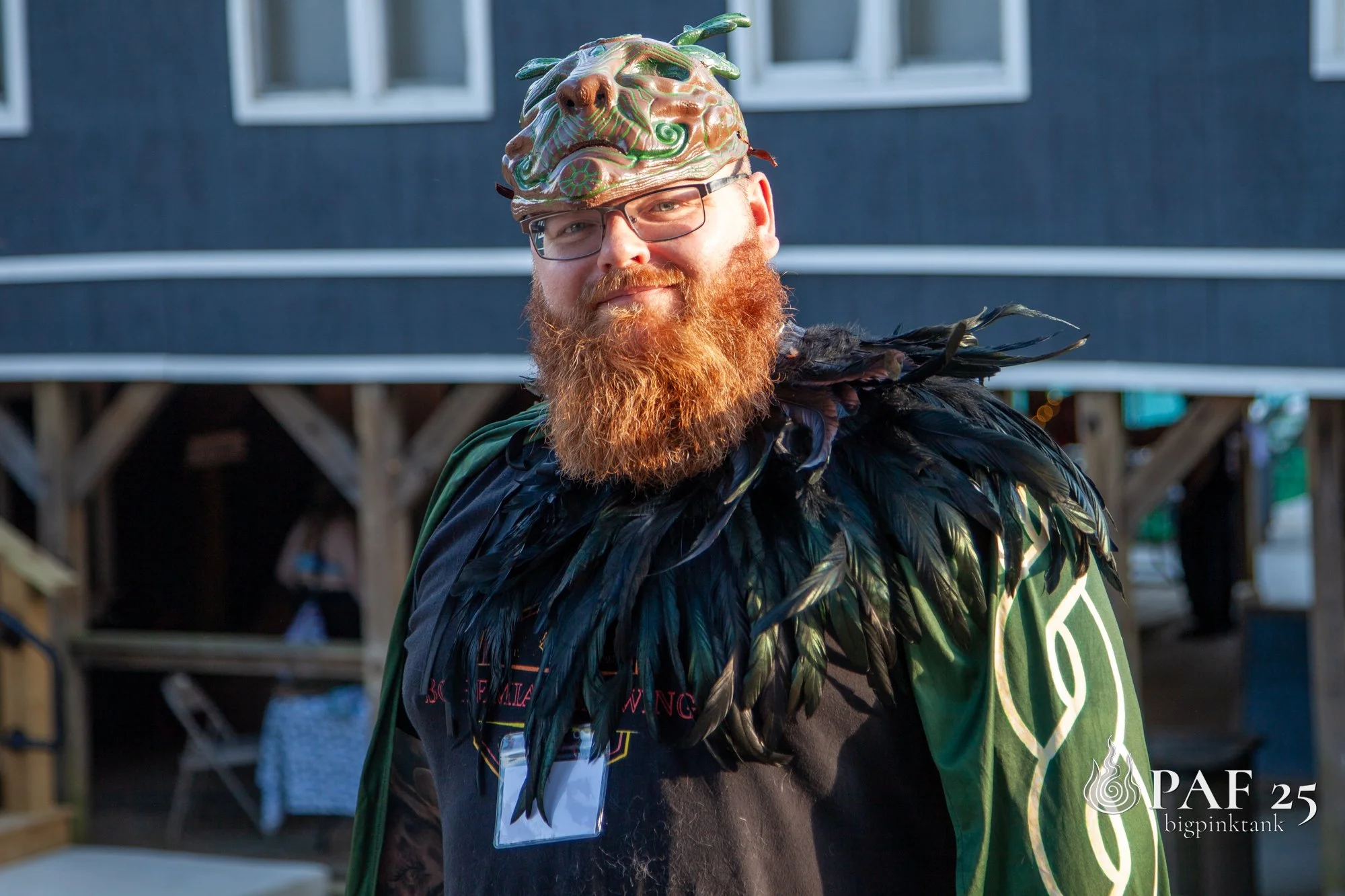 Man with red beard and glasses wearing a feathered costume and a mask resembling a lion's face, outdoors with a dark building in the background.
