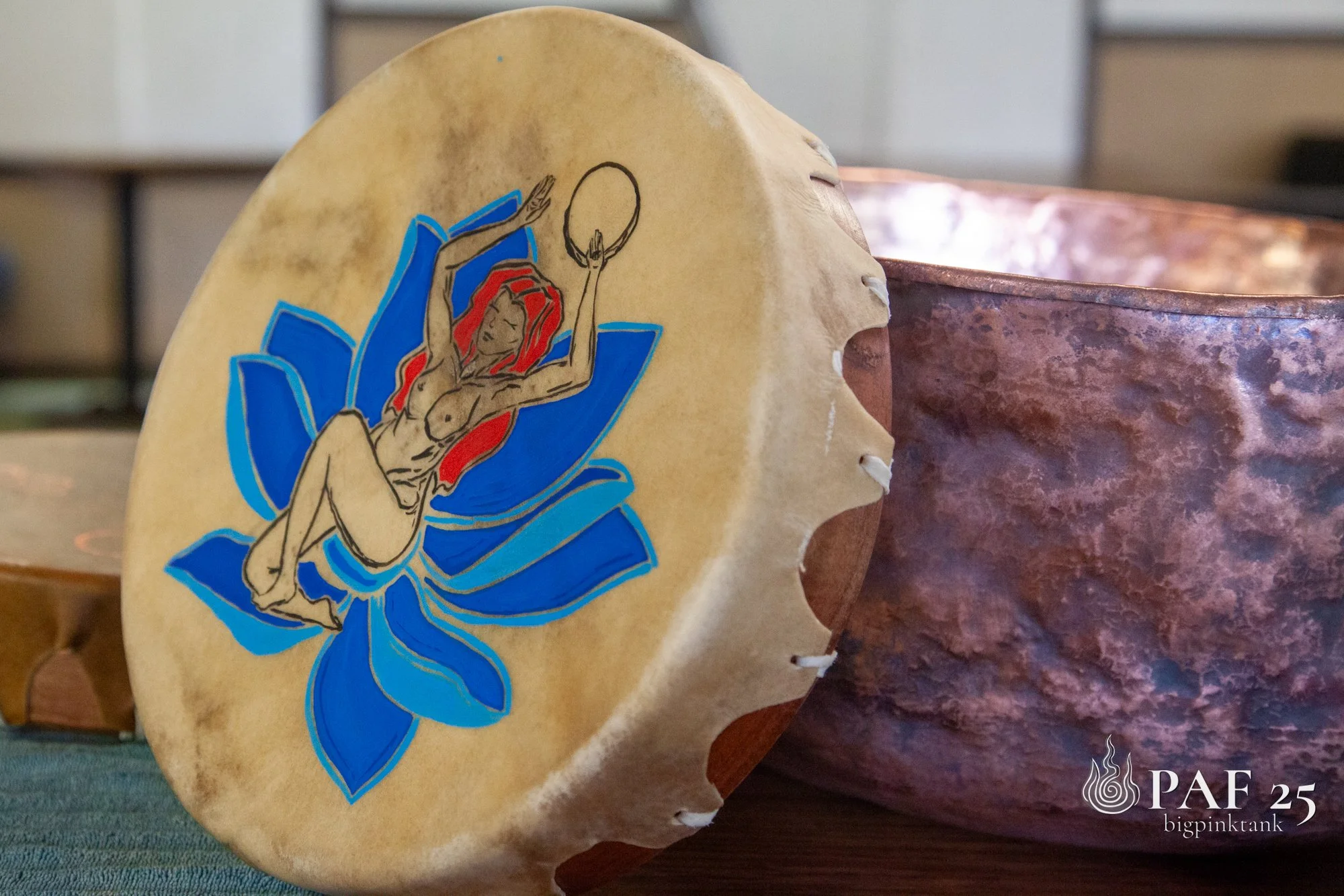 Decorative drum with a colorful illustration of a woman with red hair, surrounded by a blue lotus flower, placed on a wooden surface next to a metallic vessel.
