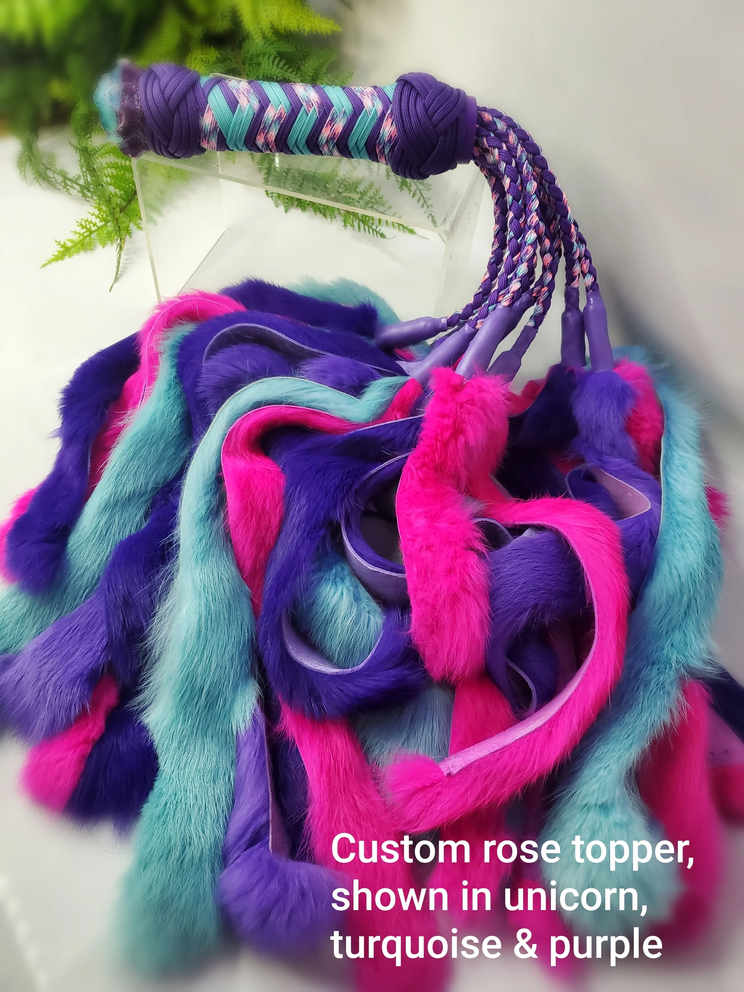 A colorful custom unicorn topper with turquoise, purple, and pink plush fur, placed on a white surface with green foliage in the background.