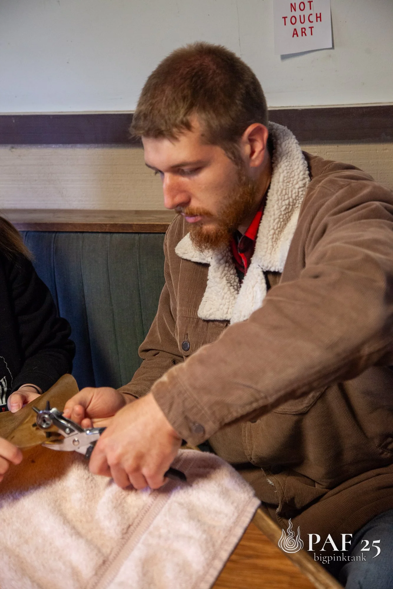 A man with a beard and short brown hair, wearing a brown jacket with a sherpa collar, is seen opening a zipper on a beige bag with a small silver object, possibly a tool or device, in his hand. A person with dark hair wearing a black shirt is partial