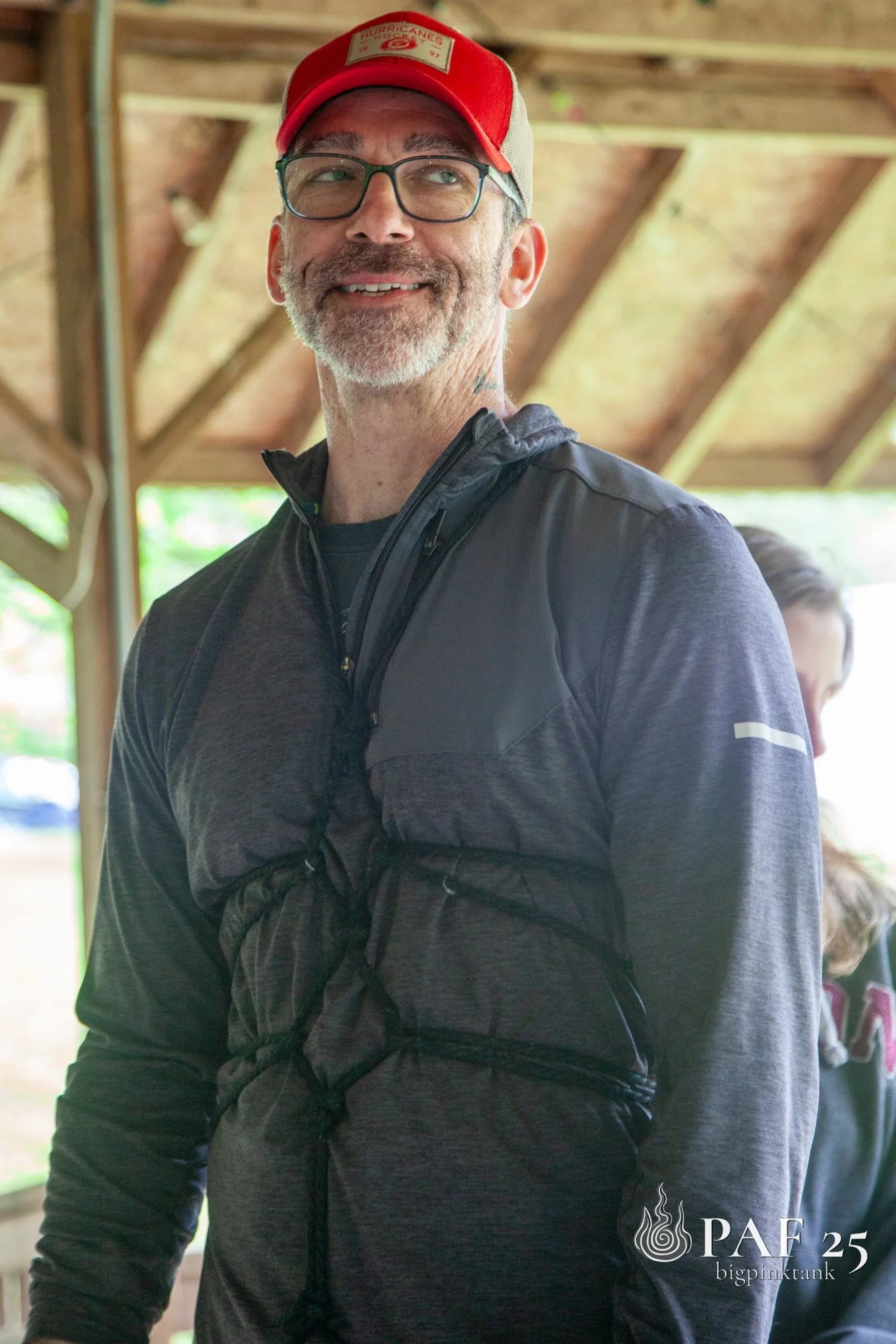 A man wearing glasses, a red cap, and a dark jacket, in a simple bondage harness , smiling indoors with a wooden roof structure and a woman partially visible in the background.
