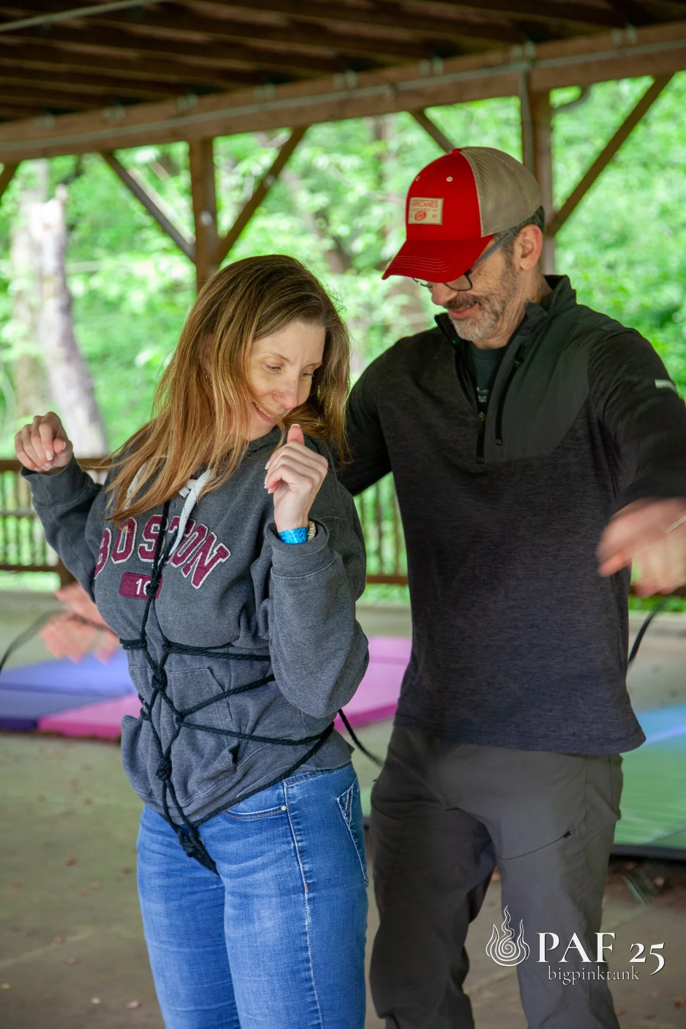 A woman and man tying shibari together under a wooden shelter in a green outdoor setting, with colorful mats on the ground.