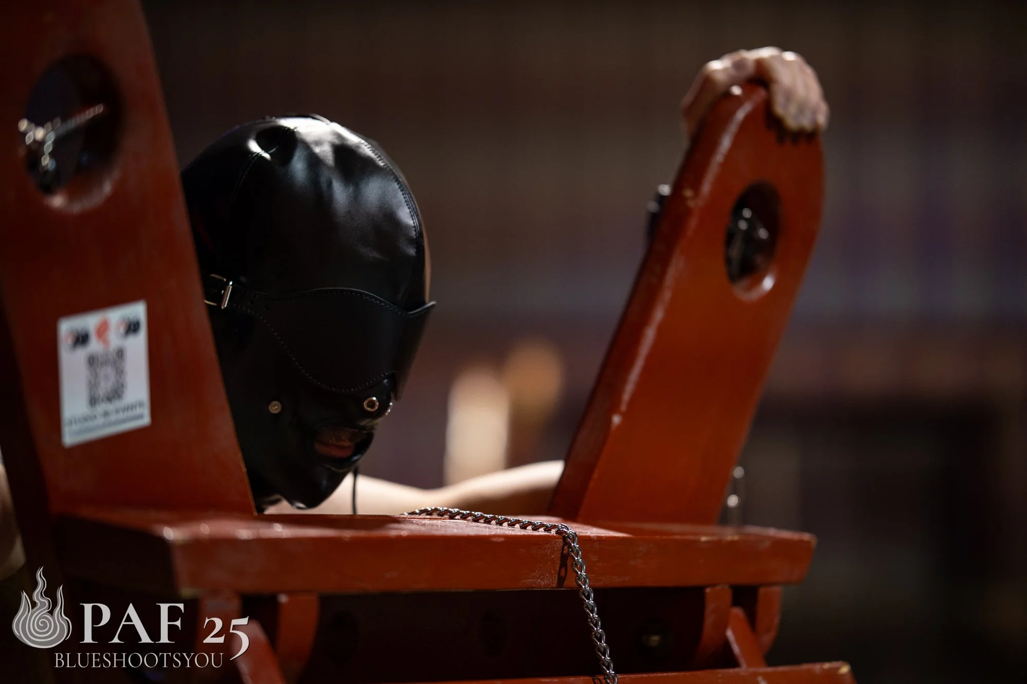 A person wears a black BDSM hood and leans against a restraint platform.