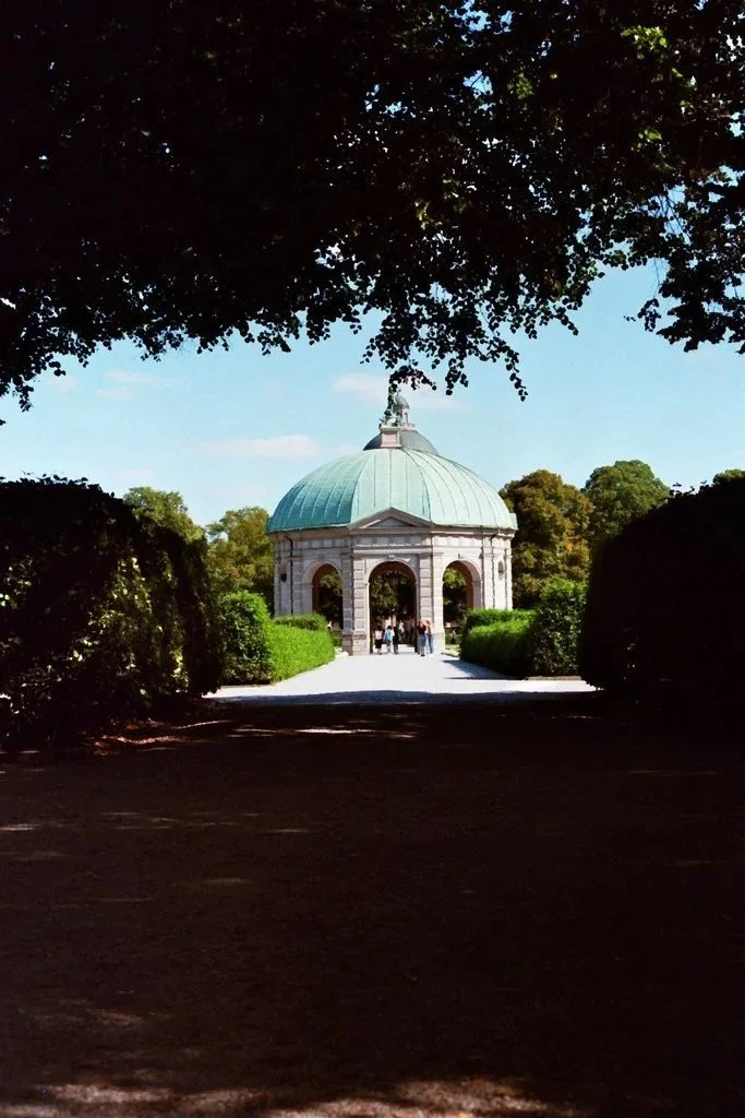 Ein Pavillon mit einer kupfergrünen Kuppel, umgeben von Büschen und Bäumen, im Freien bei sonnigem Wetter, im Hintergrund ist ein blauer Himmel mit wenigen Wolken, Besucher sind unterwegs zum Pavillon.