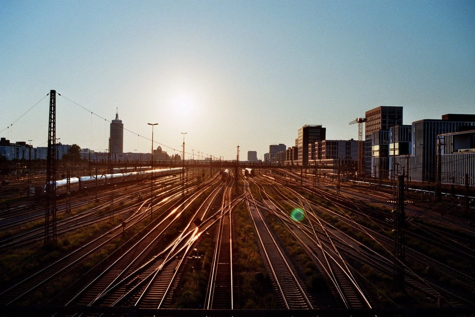 Stadtansicht mit Eisenbahnschienen bei Sonnenuntergang, moderne Gebäude im Hintergrund, einige Züge auf den Gleisen.