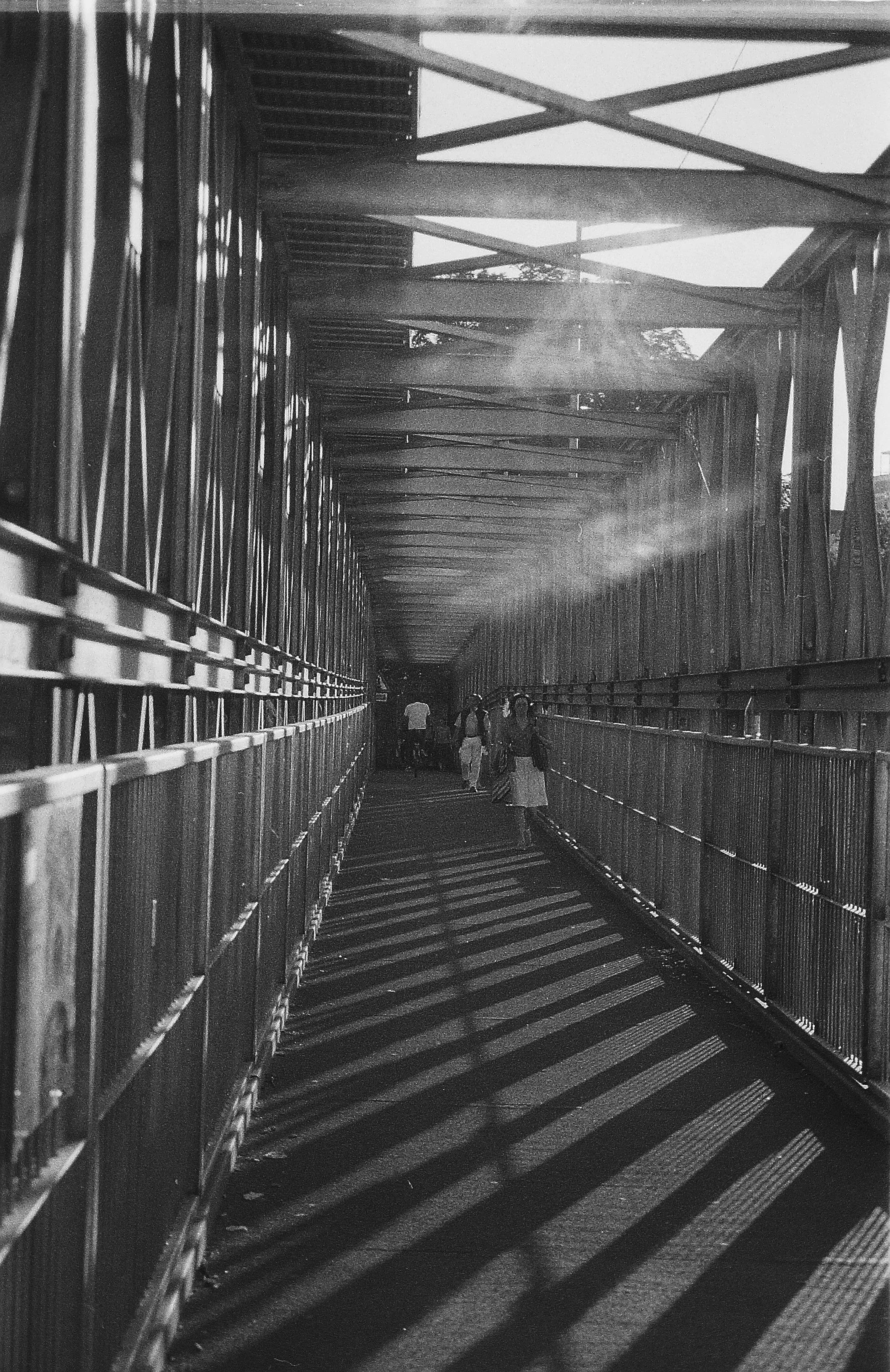 Menschen gehen durch eine Brücke mit metallicem Gitter und Stützstreben, Schatten fallen auf den Weg.