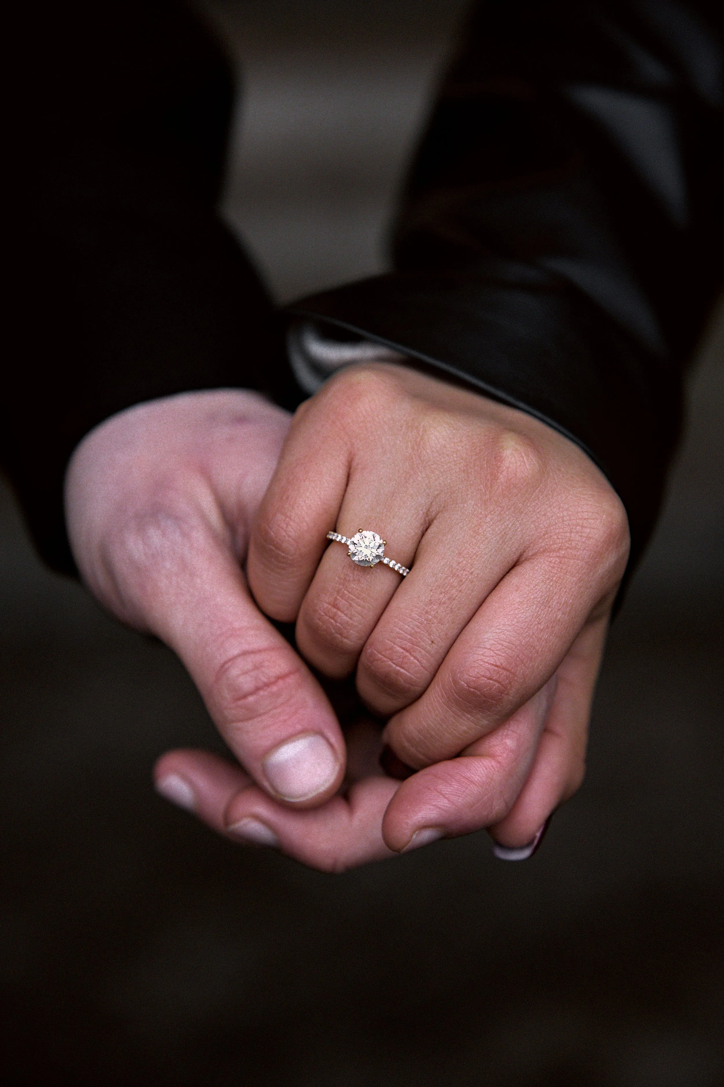Nahaufnahme von zwei Händen, die sich halten. Eine Hand trägt einen Verlobungsring mit großem Diamanten. Der Hintergrund ist dunkel, und es ist ein elegantes Kleidungsstück sichtbar.