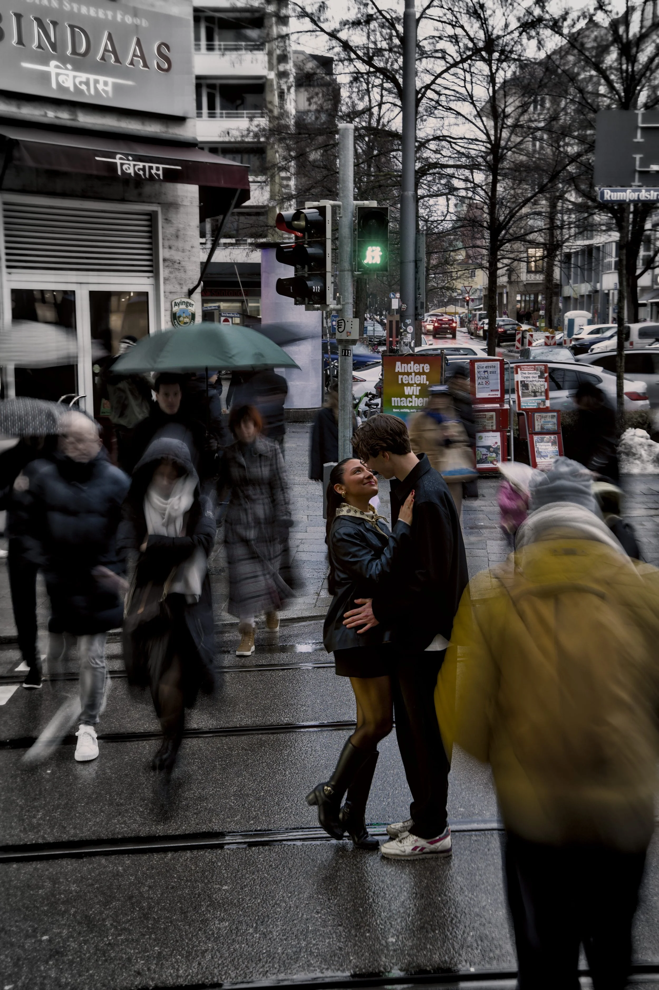 Straßenszene mit vielen Passanten bei Regen, ein Mann und eine Frau in der Mitte, die sich zu küssen scheinen, und ein grünes Fußgängersignal, im Hintergrund Geschäfte, Bäume und parkende Autos.
