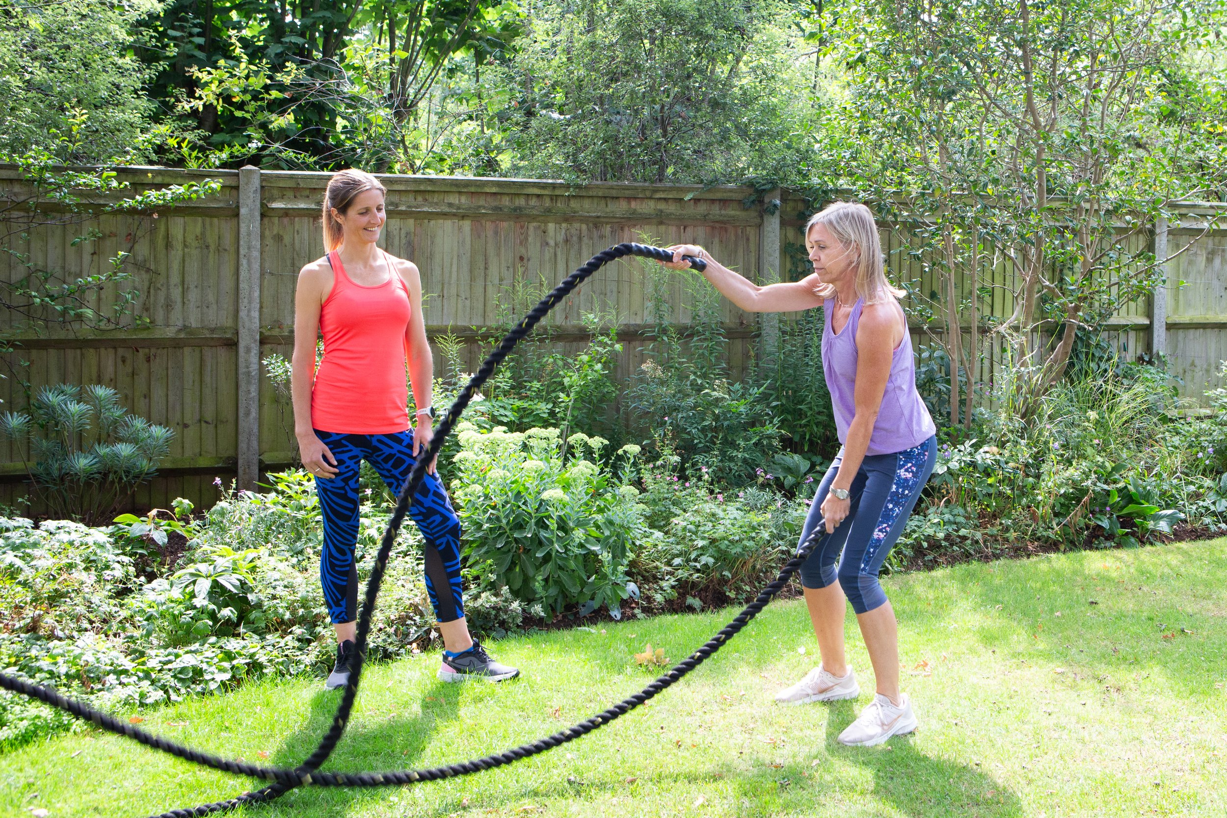 Two women exercising outdoors with battle ropes in a garden surrounded by greenery and a wooden fence.