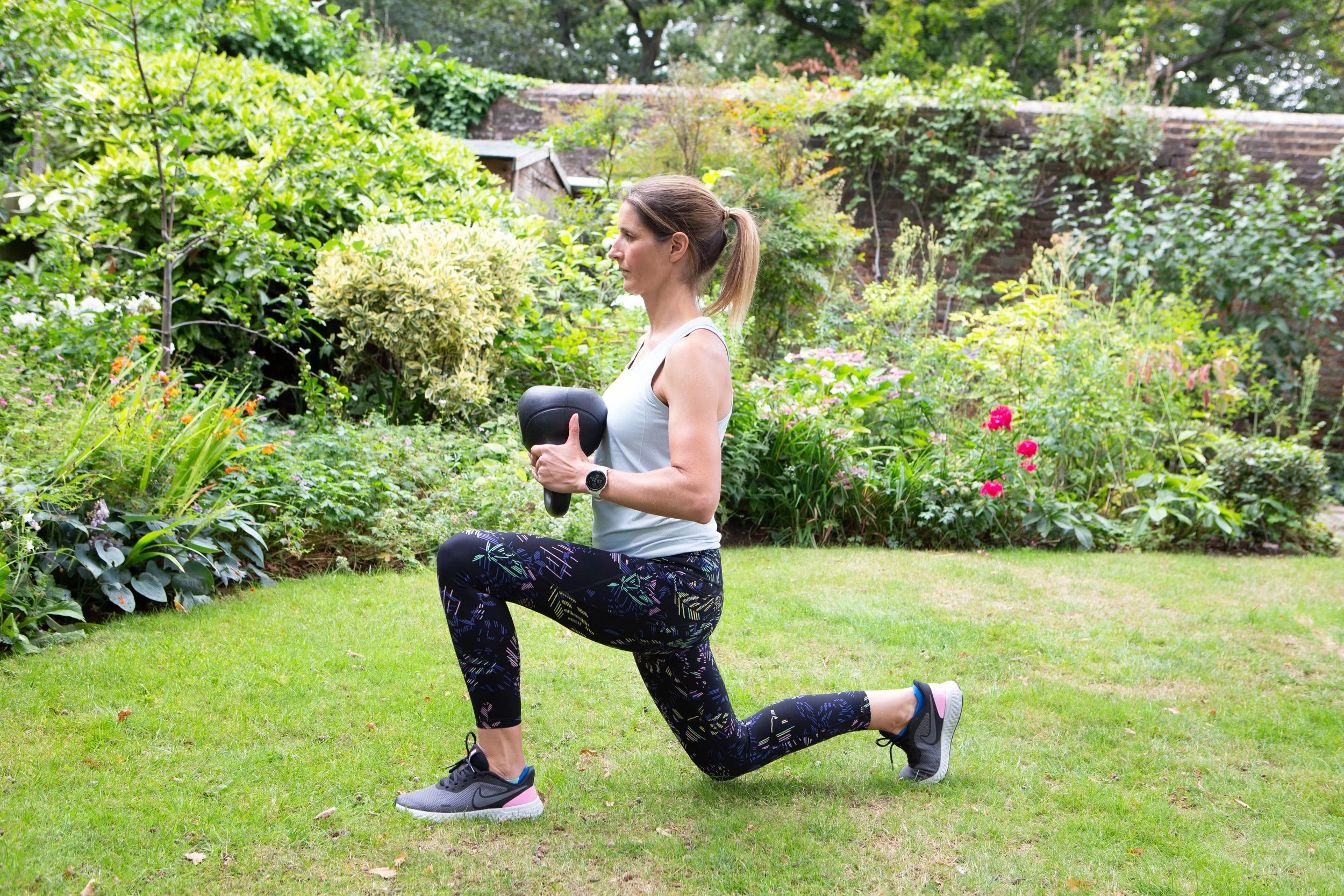 A woman performing a lunge exercise on a grassy lawn in a garden, holding a kettlebell in her hands.