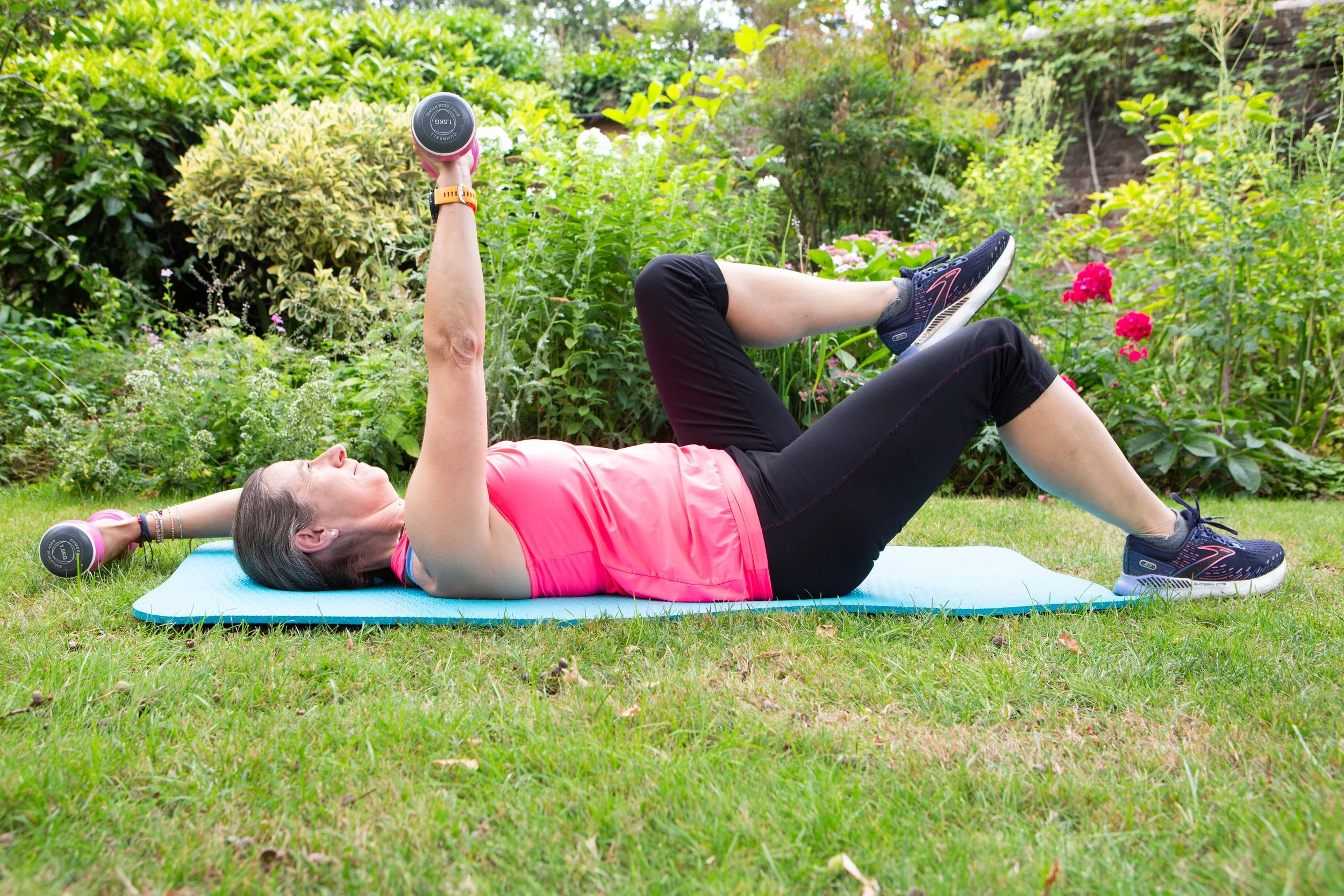 A woman exercising outdoors on a yoga mat, lying on her back with one knee bent and the other leg raised, holding dumbbells in both hands over her chest, in a garden with green bushes and flowers.