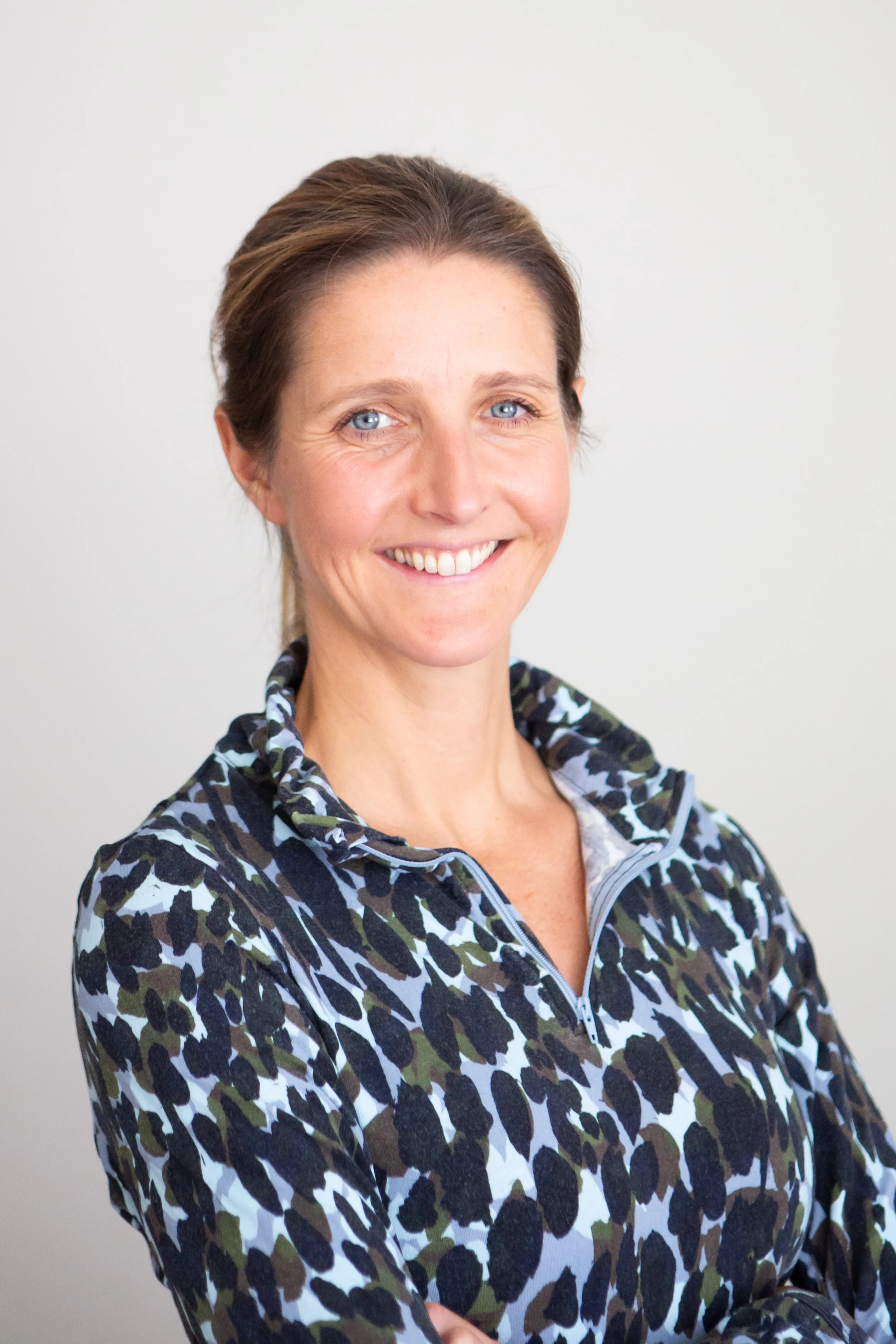 A woman with blue eyes and brown hair smiling at the camera, wearing a patterned zip-up top.