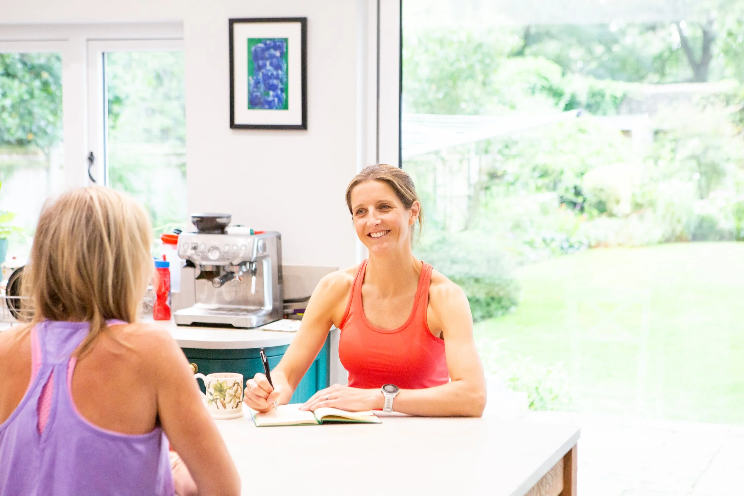 A woman in an orange tank top smiling while taking notes at a white kitchen table during a conversation with a girl with blonde hair in a purple top.