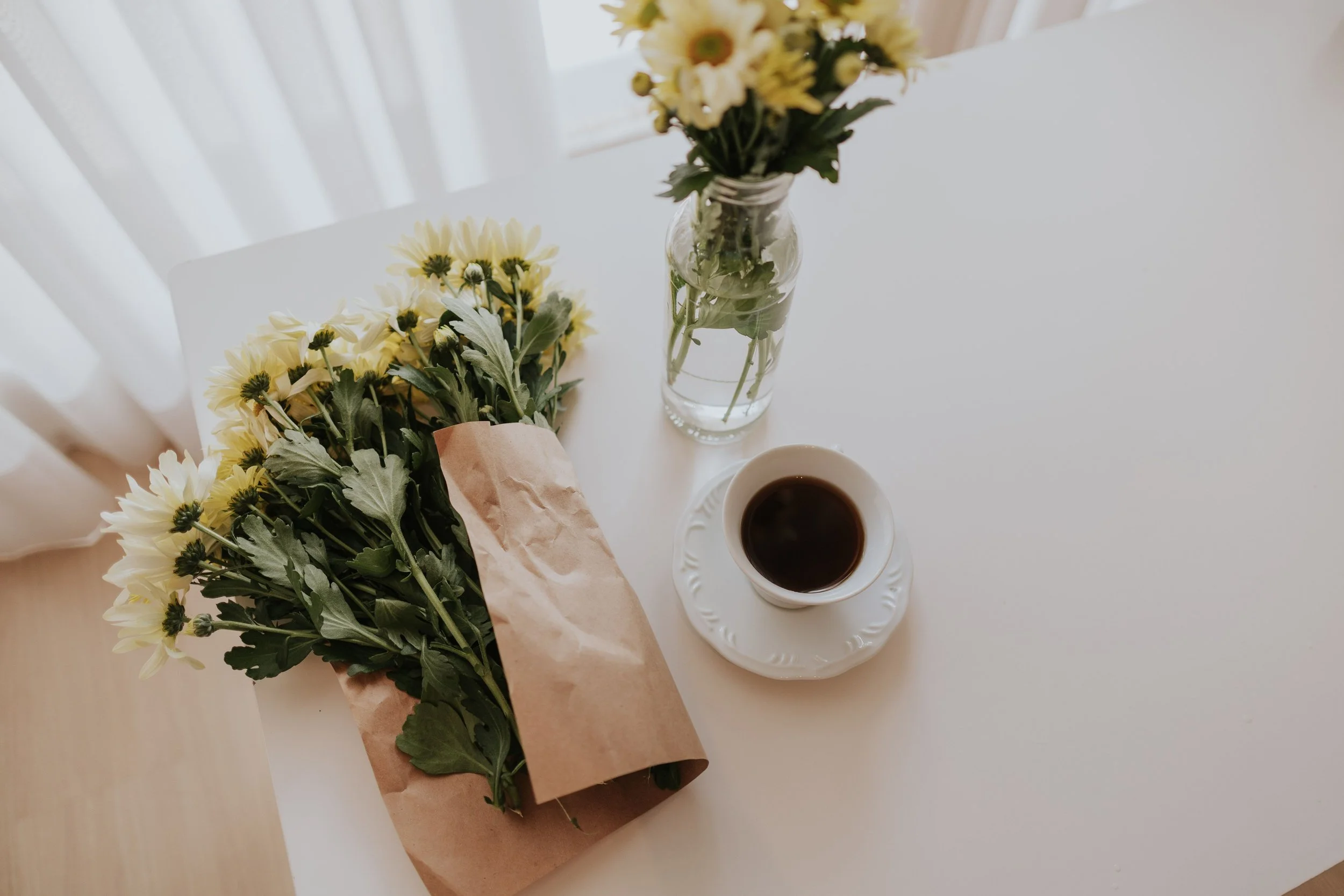 A white table holds a bouquet of yellow and white flowers in a glass jar, a matching smaller bouquet wrapped in brown paper, a cup of black coffee on a white saucer, with sunlight streaming through sheer curtains in the background.