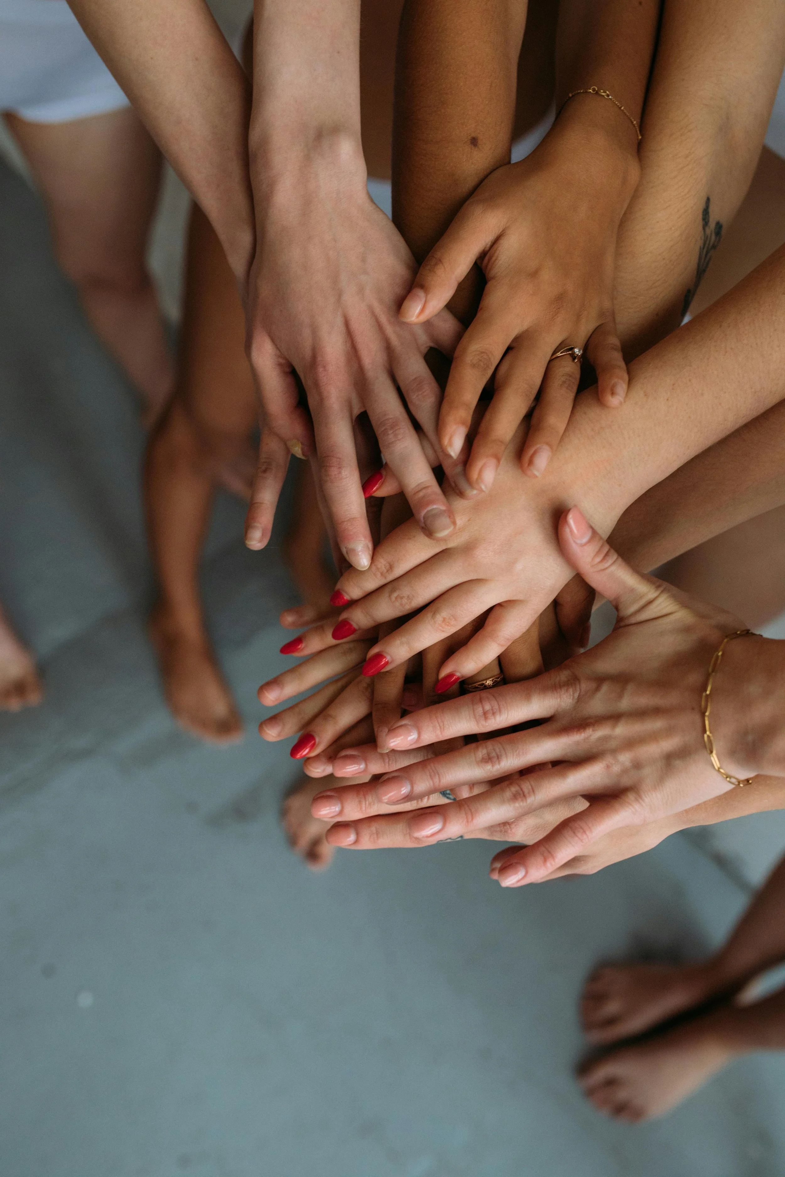 Multiple hands stacked in the center, with some wearing rings and bracelets, and a few with painted red nails, forming a display of unity and friendship.