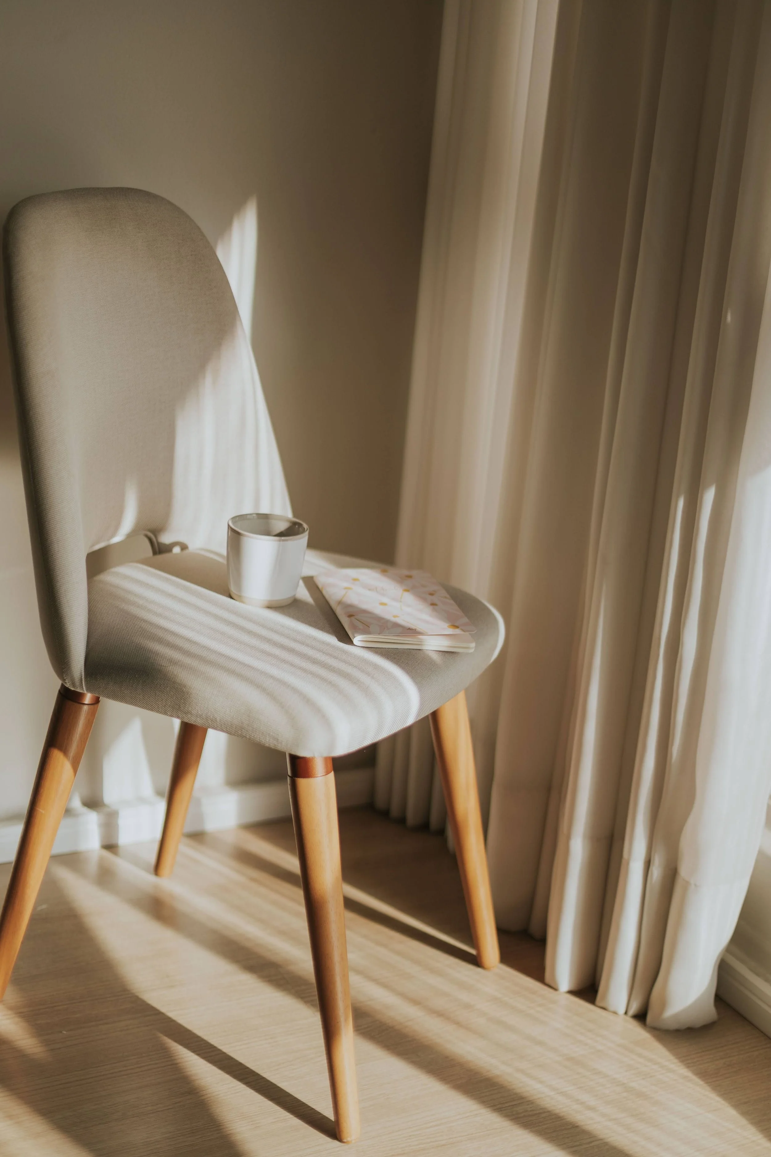 A beige upholstered chair with wooden legs, placed beside beige curtains with sunlight casting shadows.
