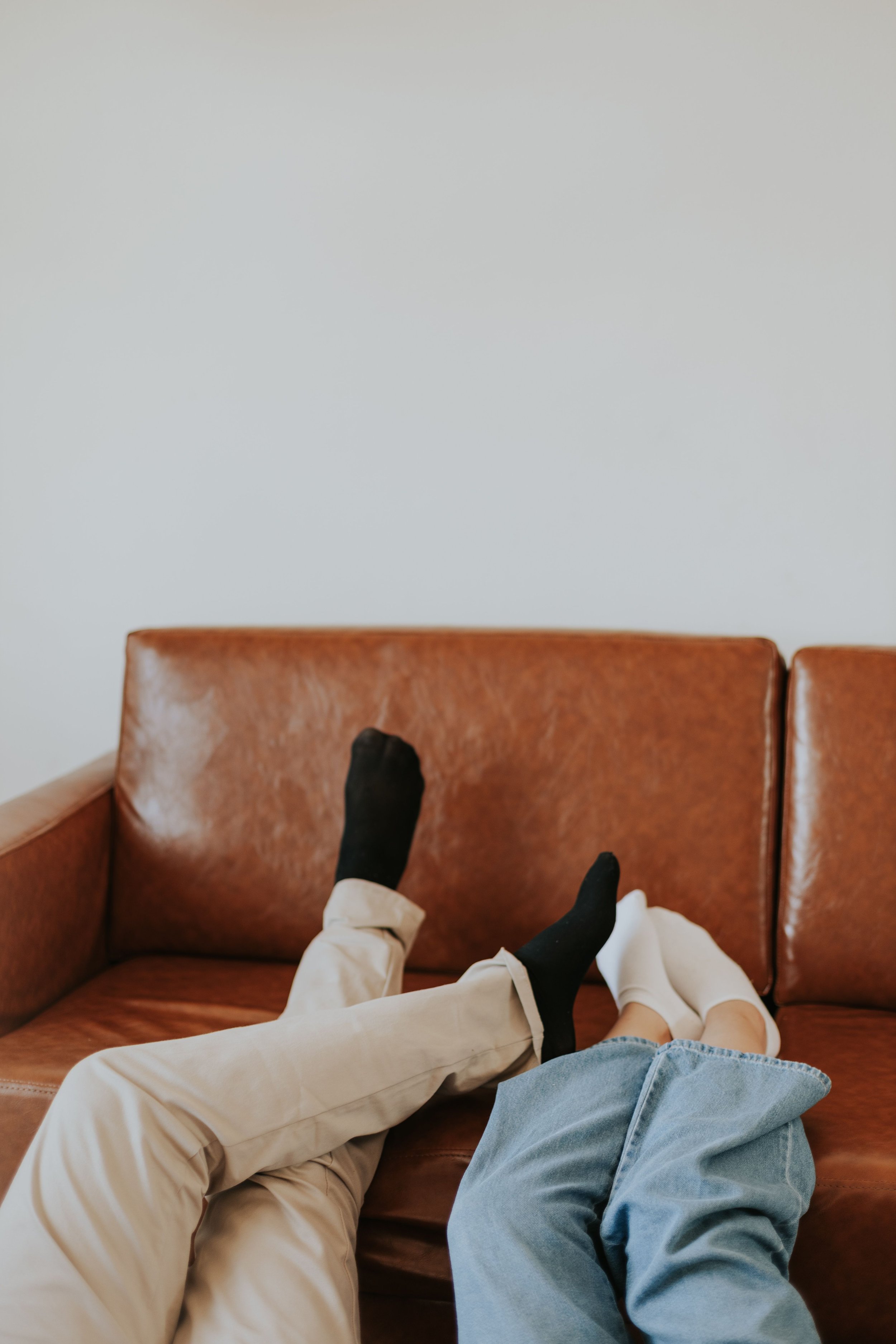 Person lying on a brown leather couch with their legs stretched out, wearing beige pants, a blue denim jacket, and black socks, against a plain light-colored wall.