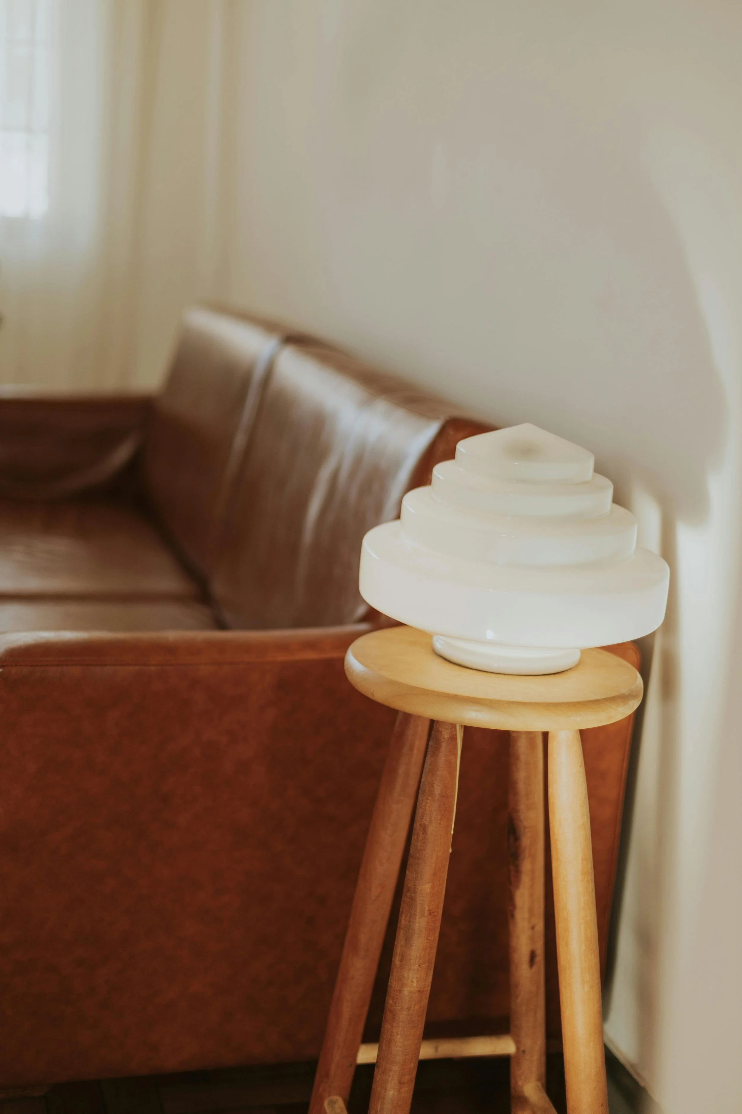 A small wooden side table holding a white layered ceramic sculpture, positioned next to a brown upholstered sofa, in a cozy room with light-colored walls.
