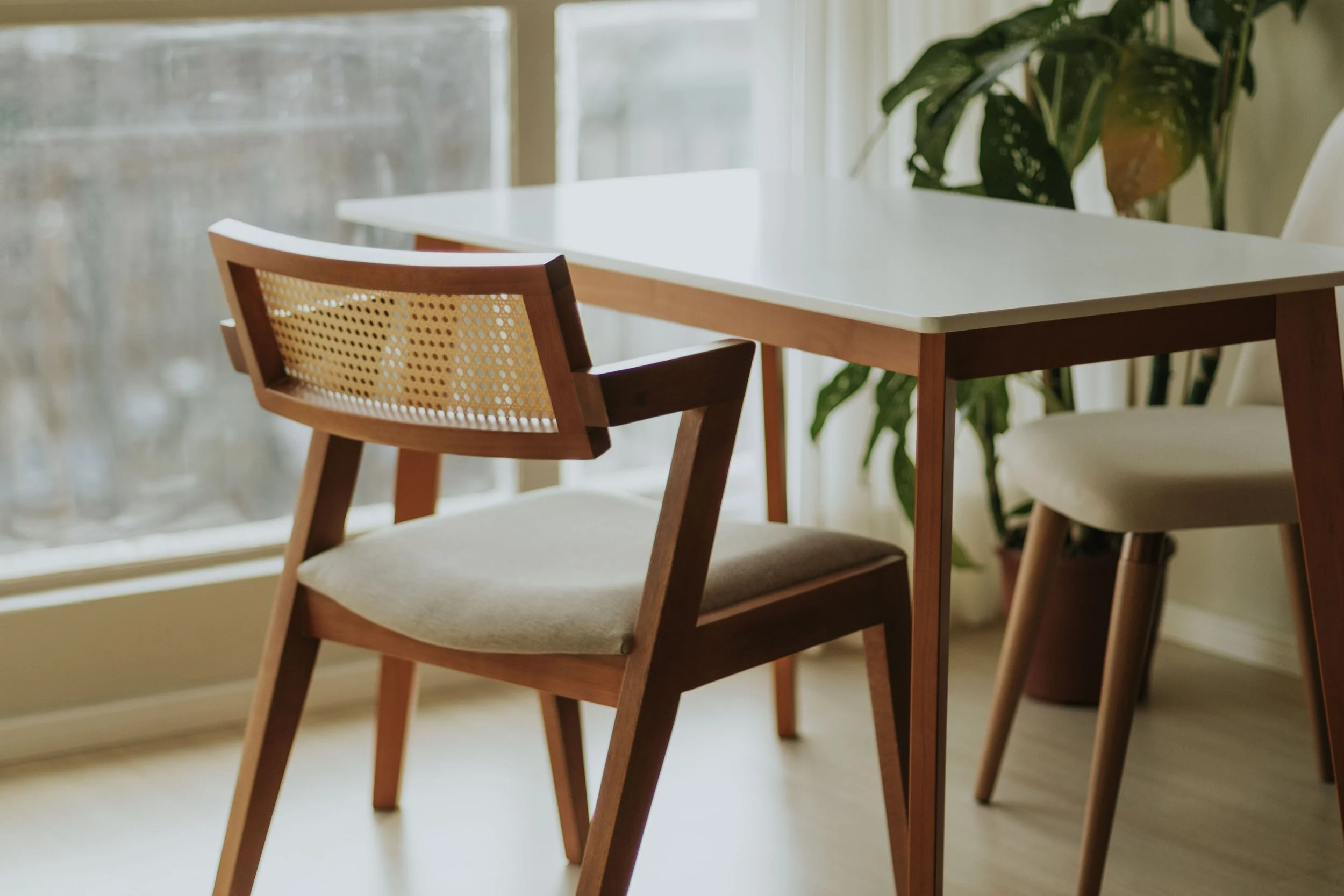 A kitchen or dining area with a wooden chair with a cane backrest and beige cushion, a white table, and a large window with a sheer curtain, with green plants nearby.