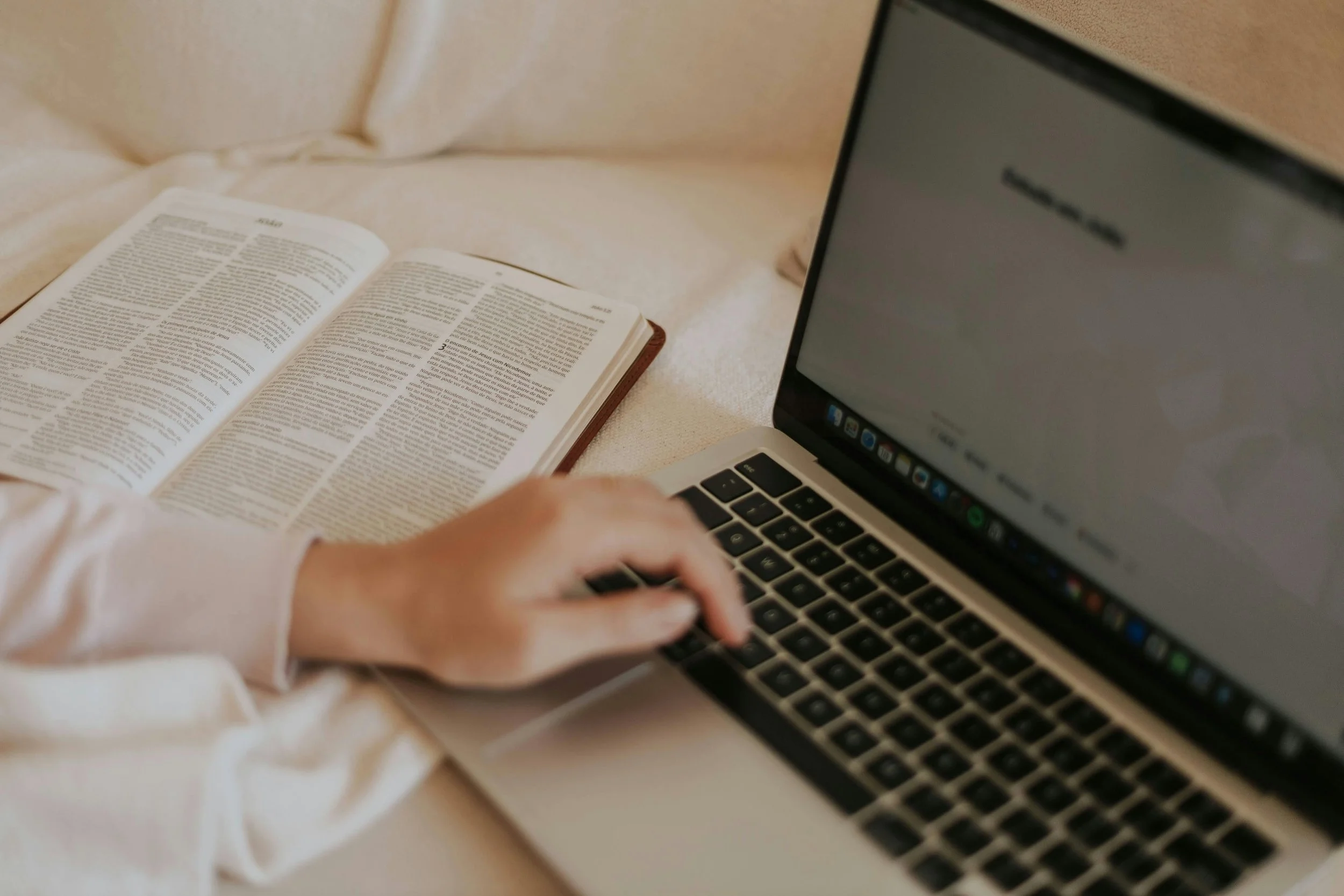 A person using a laptop with an open book resting on a soft surface nearby.