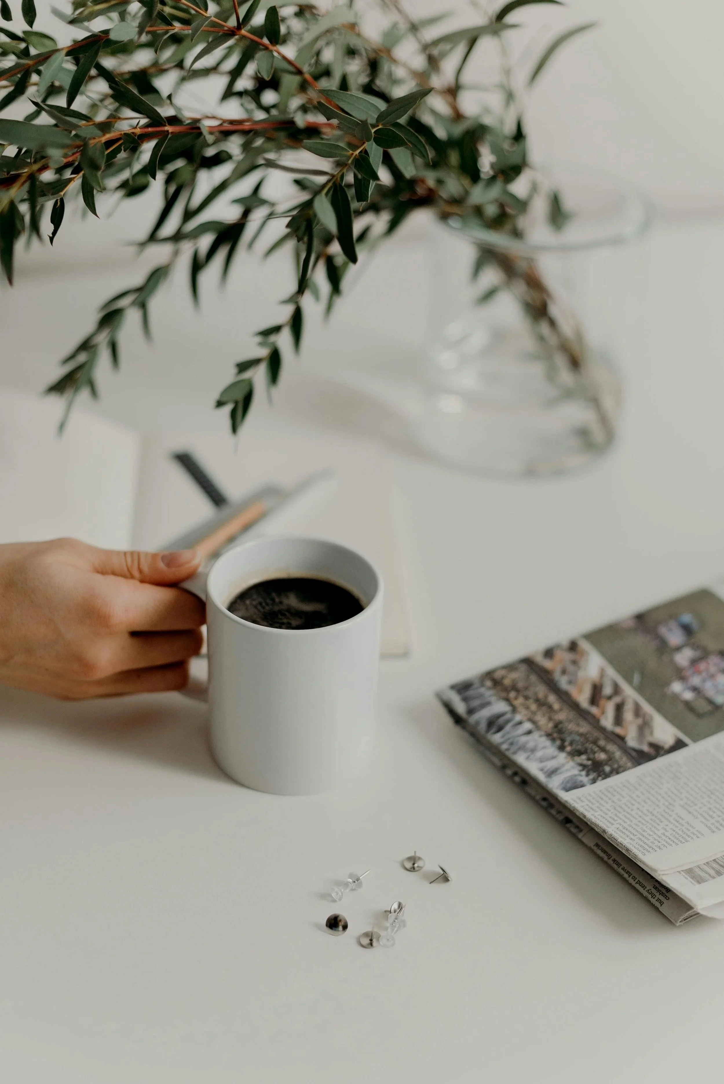 A white coffee mug filled with coffee, held by a person's hand, on a white table next to a newspaper, a few metallic push pins, and a glass vase with green leafy plant.