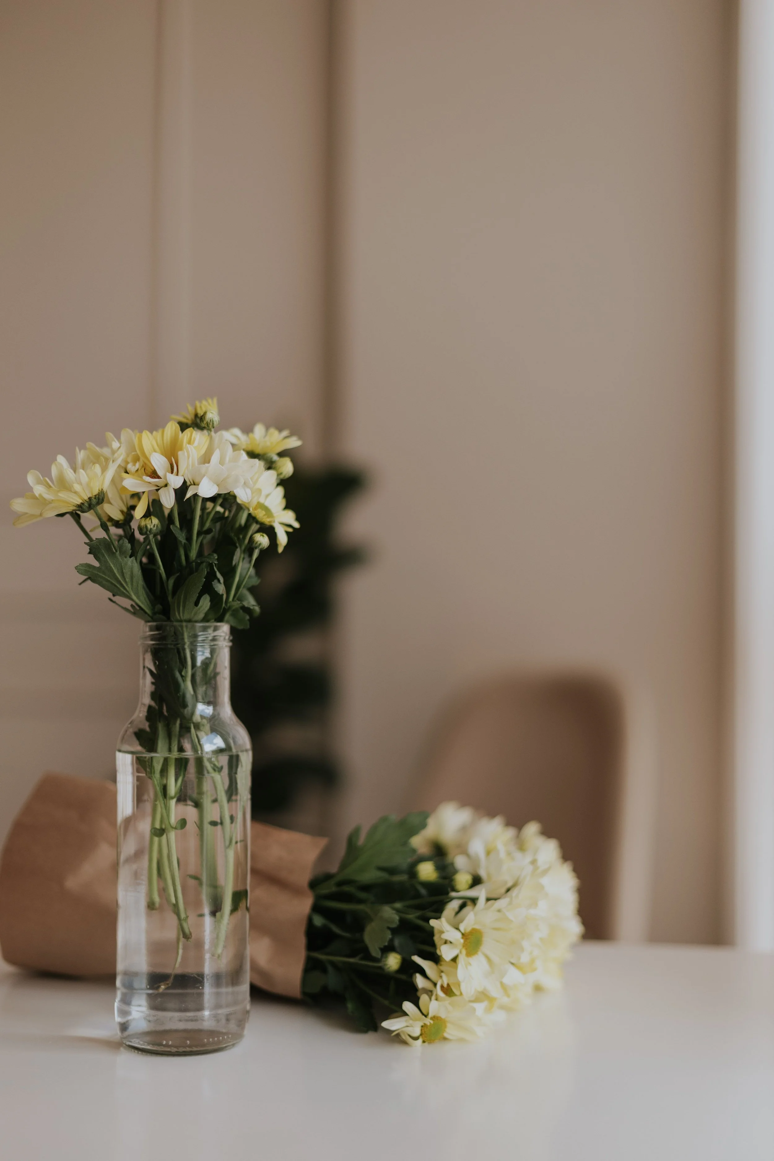 White and yellow flowers in a clear glass vase on a white surface, with additional flowers and foliage in the background.