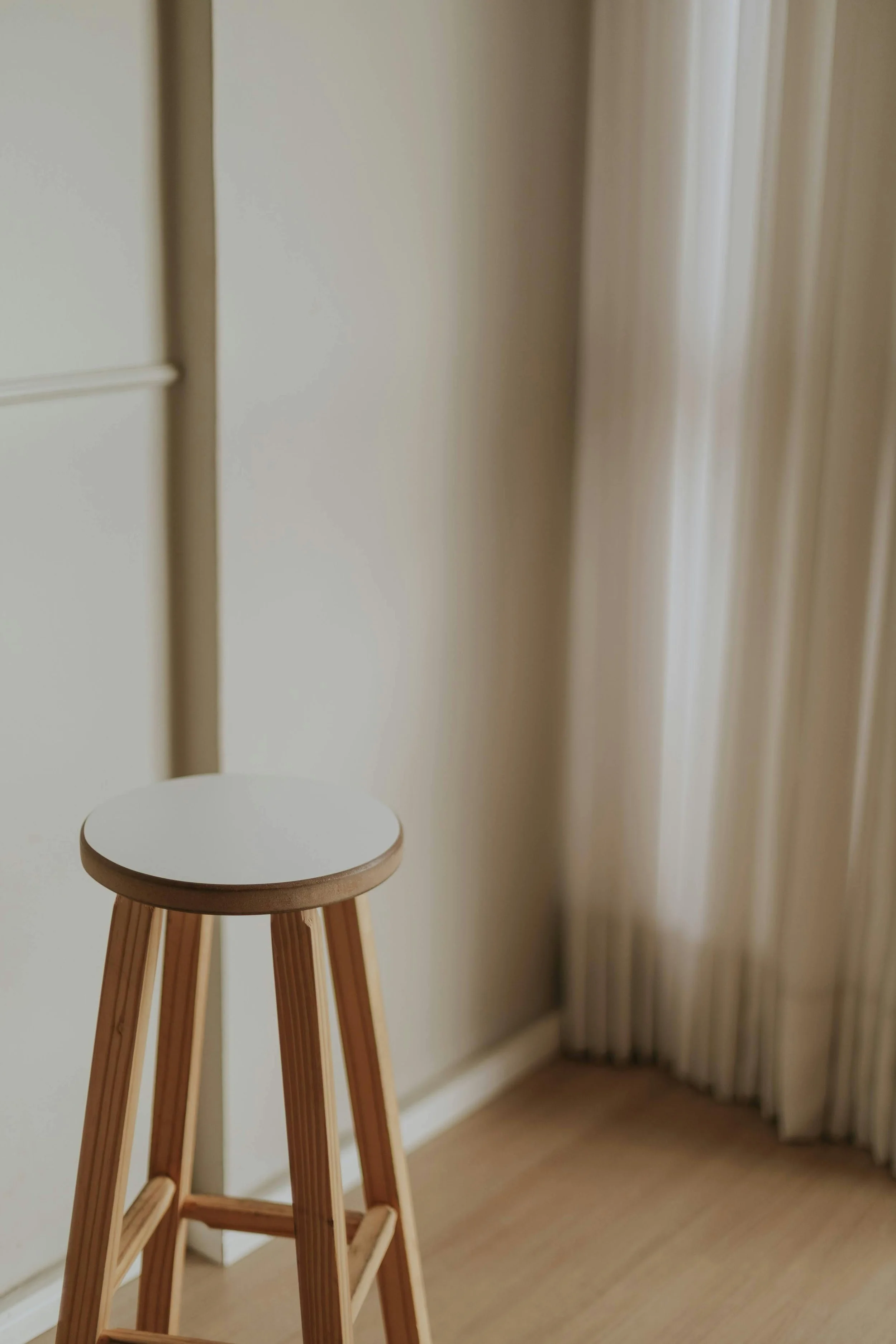 A wooden stool with a round white seat in a room with beige curtains and light-colored wooden flooring.