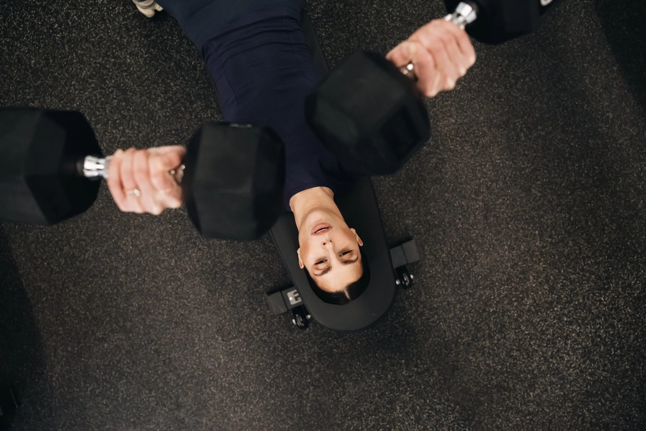 A woman lying on her back on a workout bench, lifting dumbbells in a gym setting. She is wearing a dark athletic top and pants, with a focused expression, and there is a textured gym floor around her.