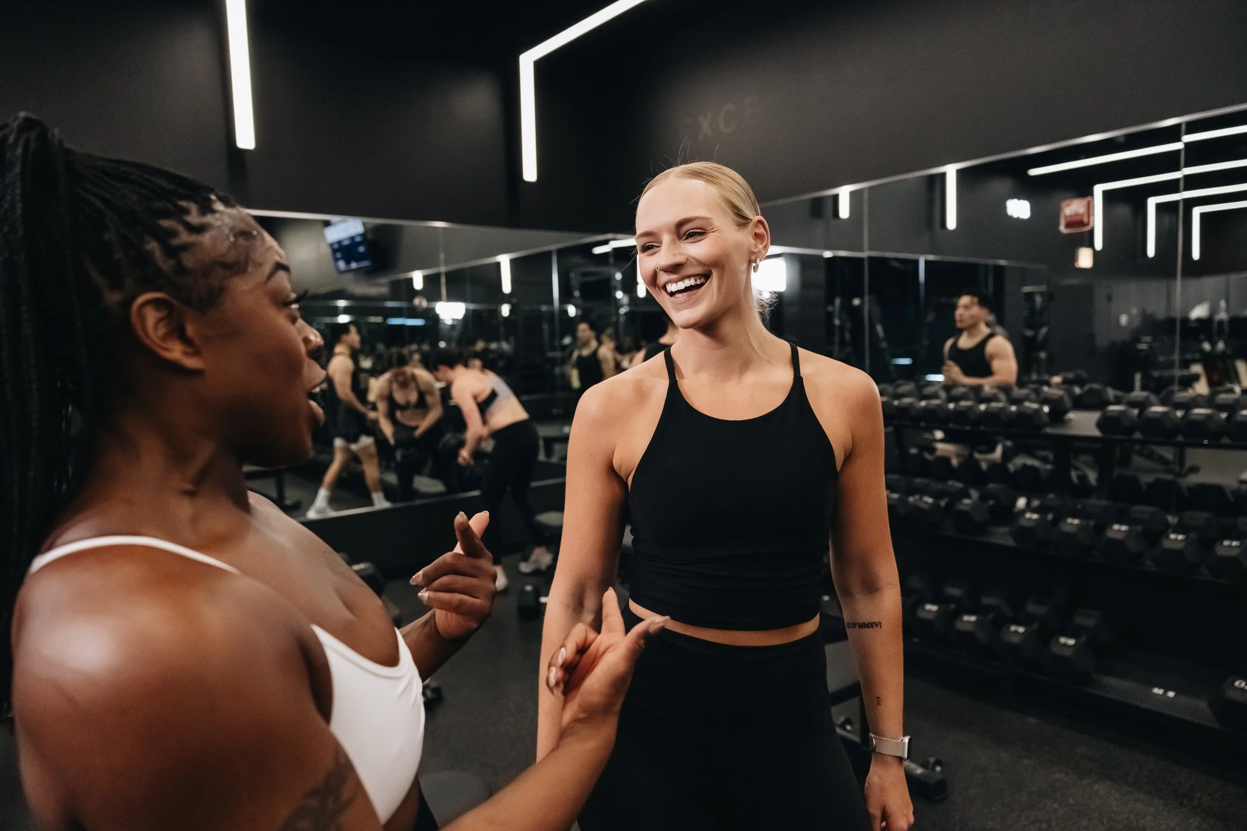 Two women talking and smiling at each other inside a gym with black walls, mirrors, and workout equipment.