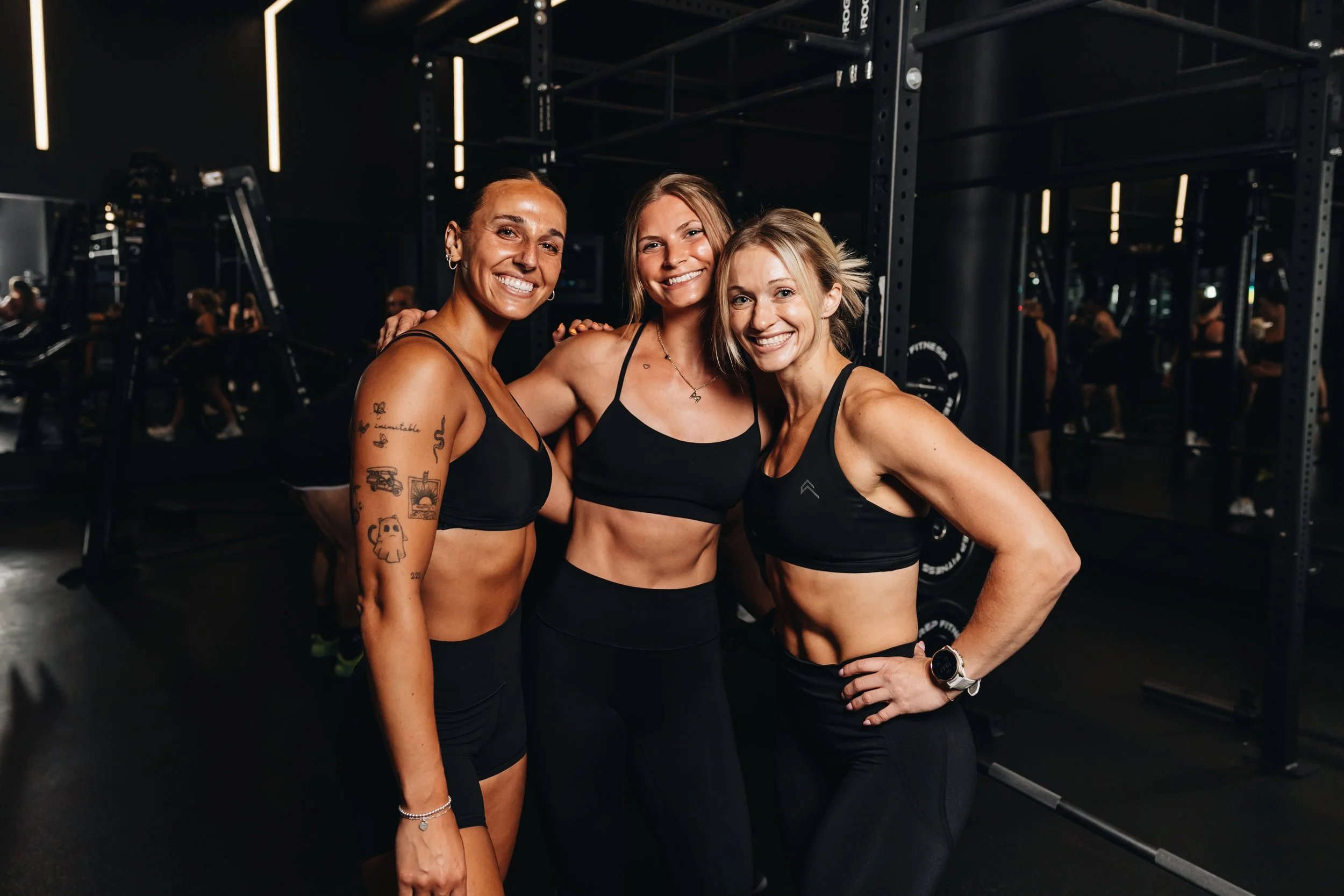 Three women in black sports bras smiling and posing together inside a gym with workout equipment in the background.