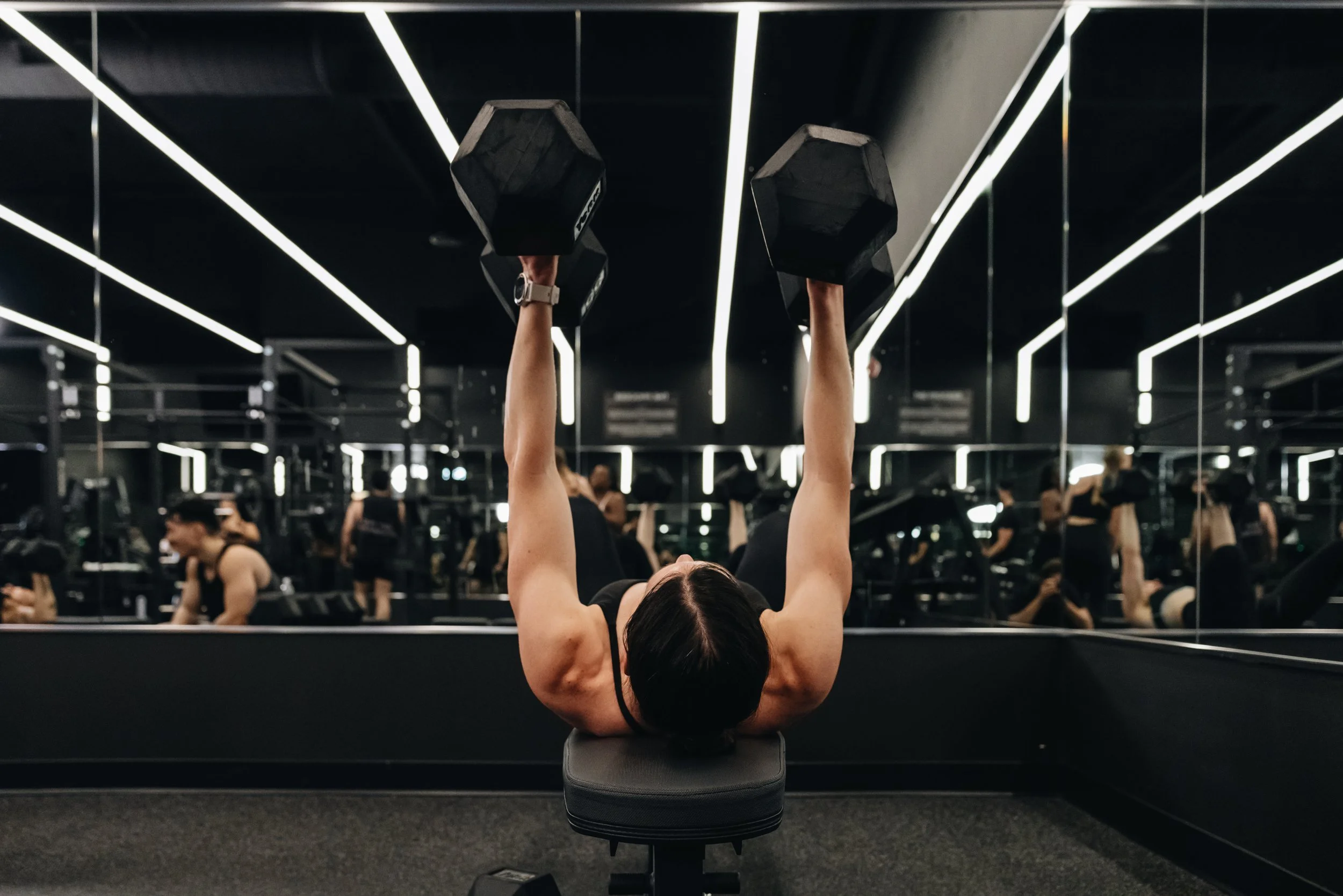 A woman lying on a workout bench lifting dumbbells in a gym with mirrored walls and people working out in the background.