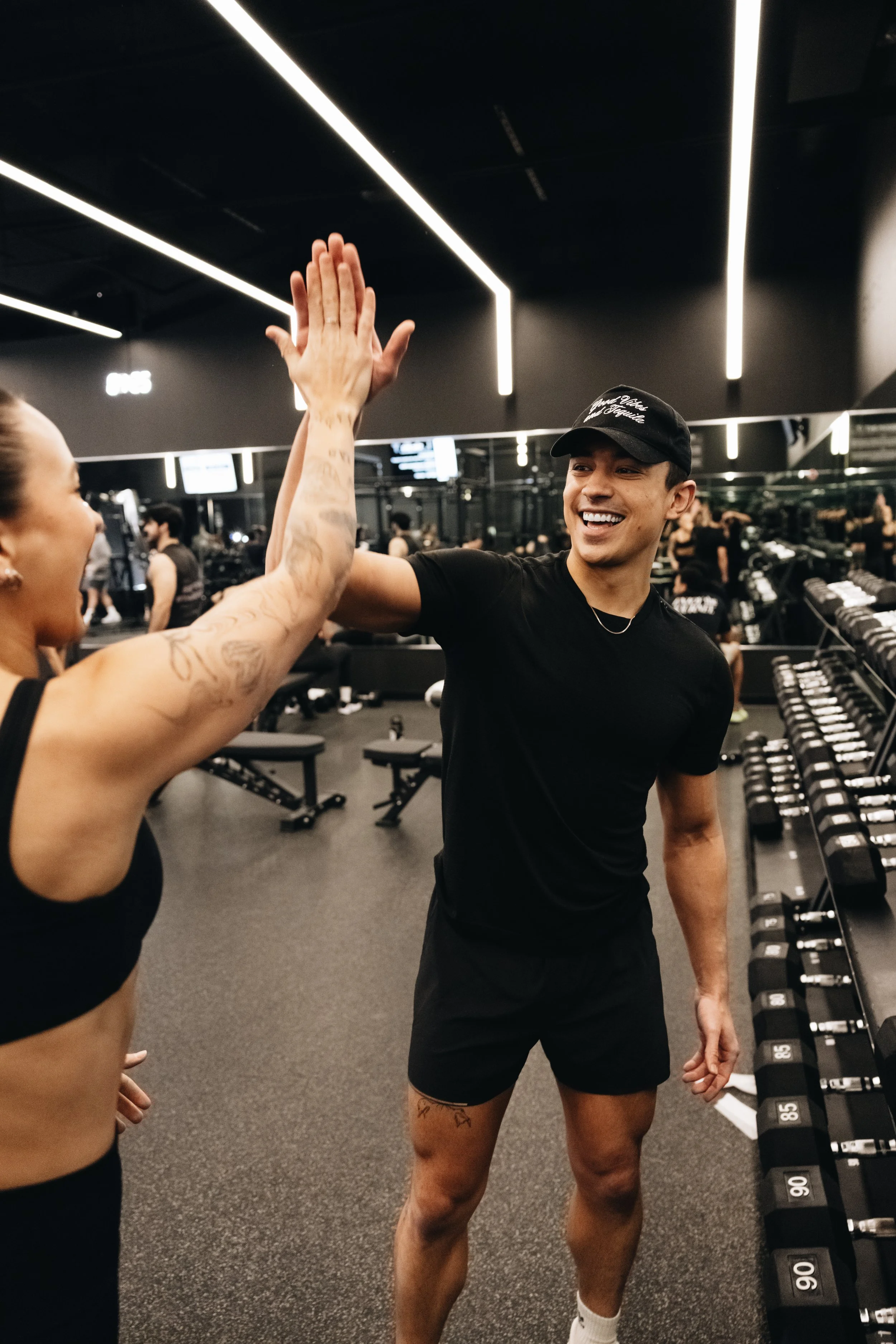 A young man and woman high-fiving in a gym with dumbbells and workout equipment in the background.