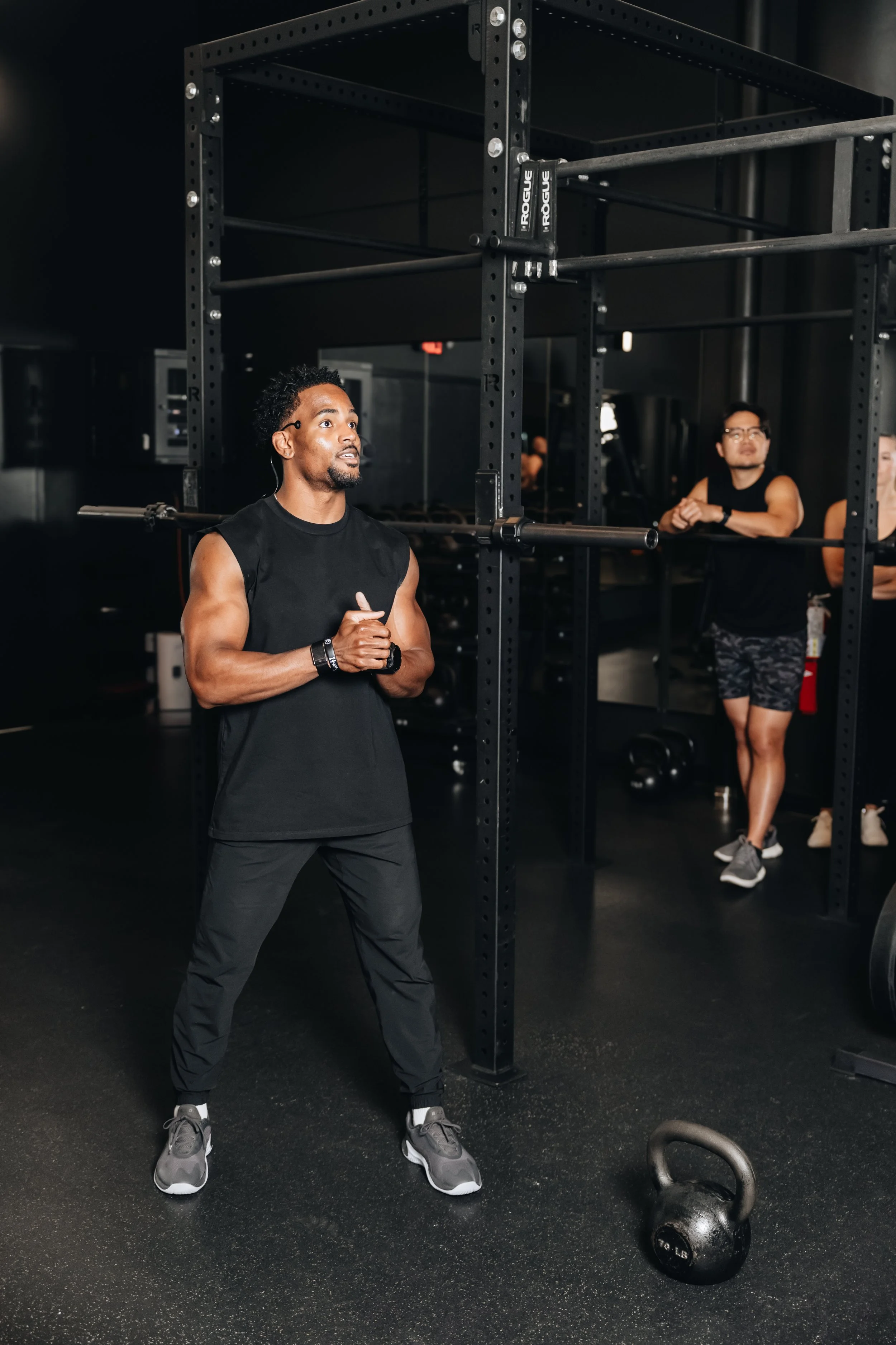 A man in black workout clothes standing in a gym near a black metal power rack, preparing to lift a barbell. Other people are seen in the background, and a kettlebell is on the floor nearby.