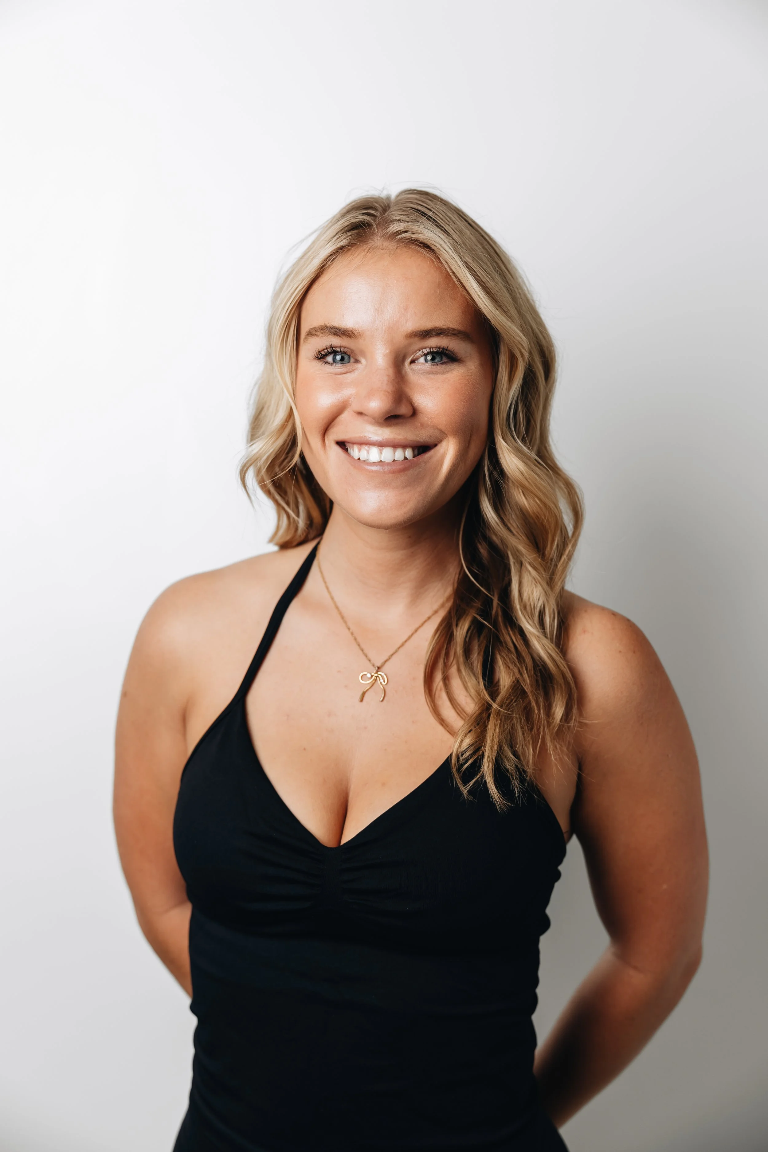 A smiling woman with long blonde hair in loose waves, wearing a black sleeveless top and a gold necklace, standing against a plain white wall.