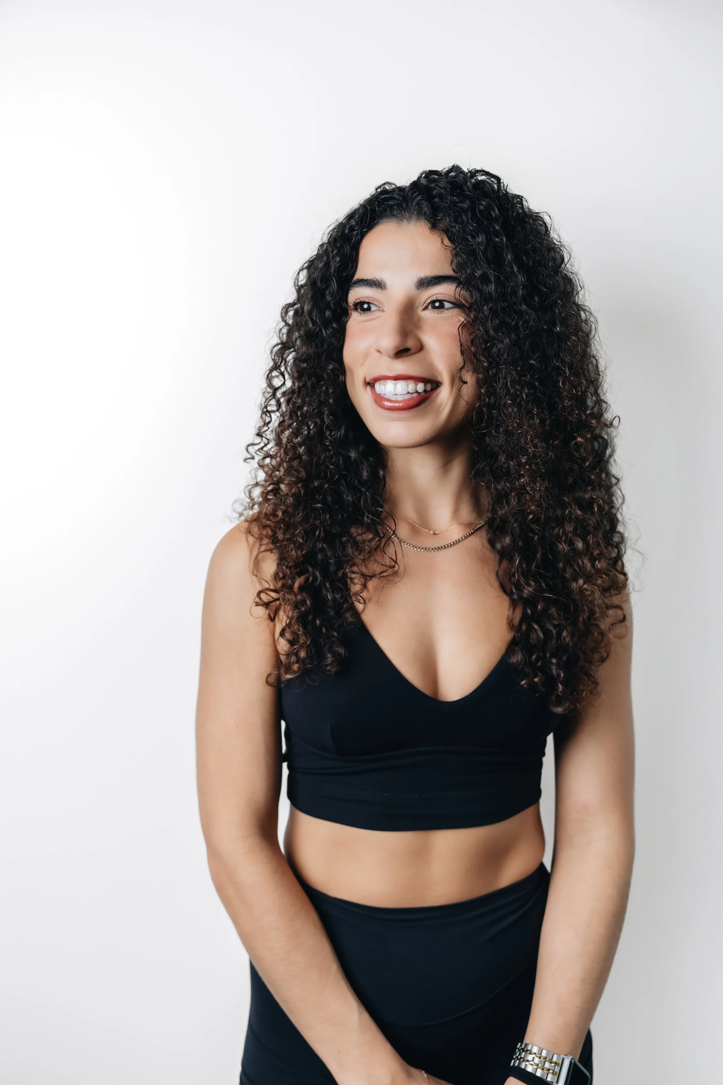 A woman with curly dark hair smiling and looking to her left, wearing a black sleeveless top and a silver watch on her left wrist, standing against a plain white background.