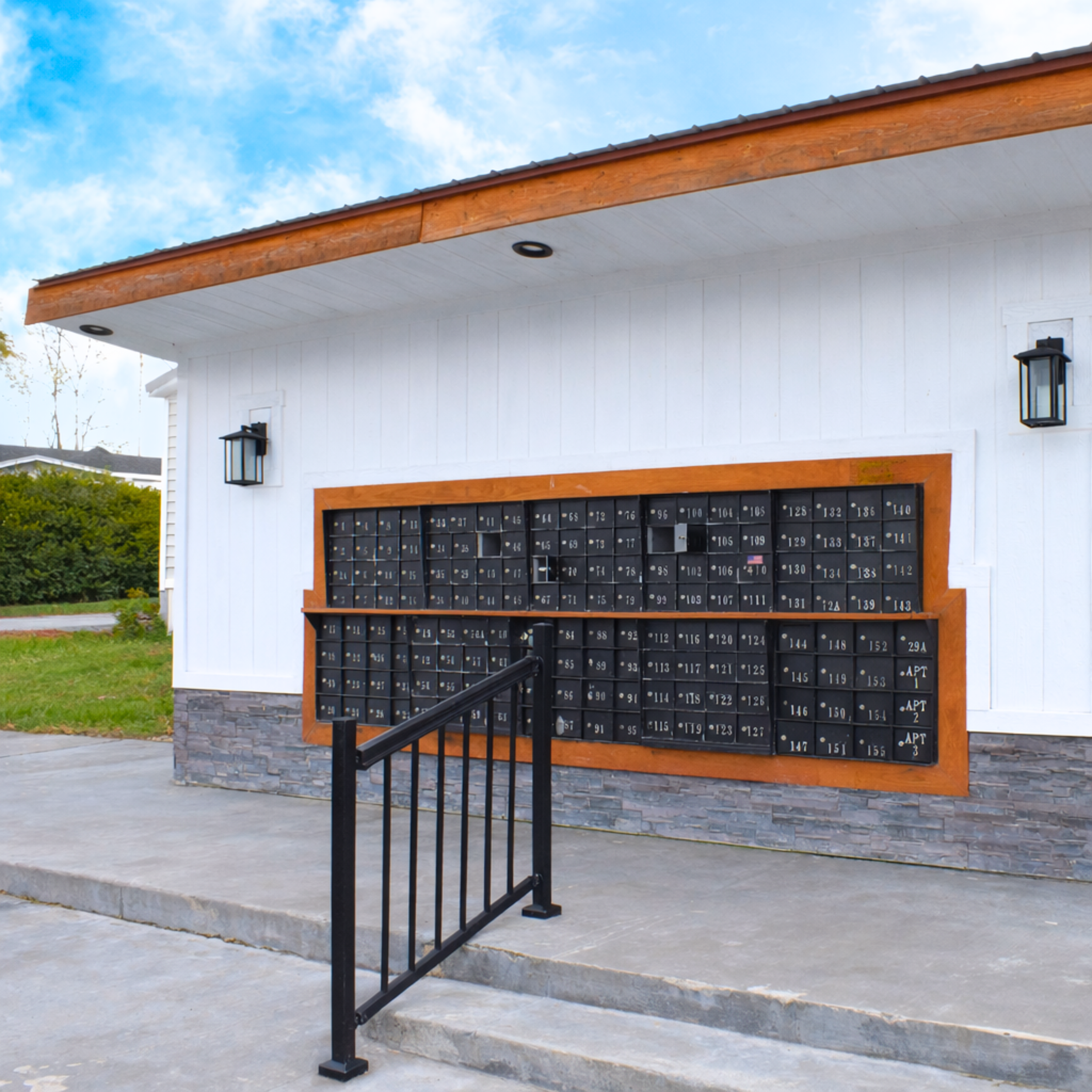 Outside view of a modern apartment mailbox area with multiple black mailboxes encased in wood, flanked by black wall-mounted lanterns, and a black metal railing, against a white building with a stone base, under a partly cloudy sky.