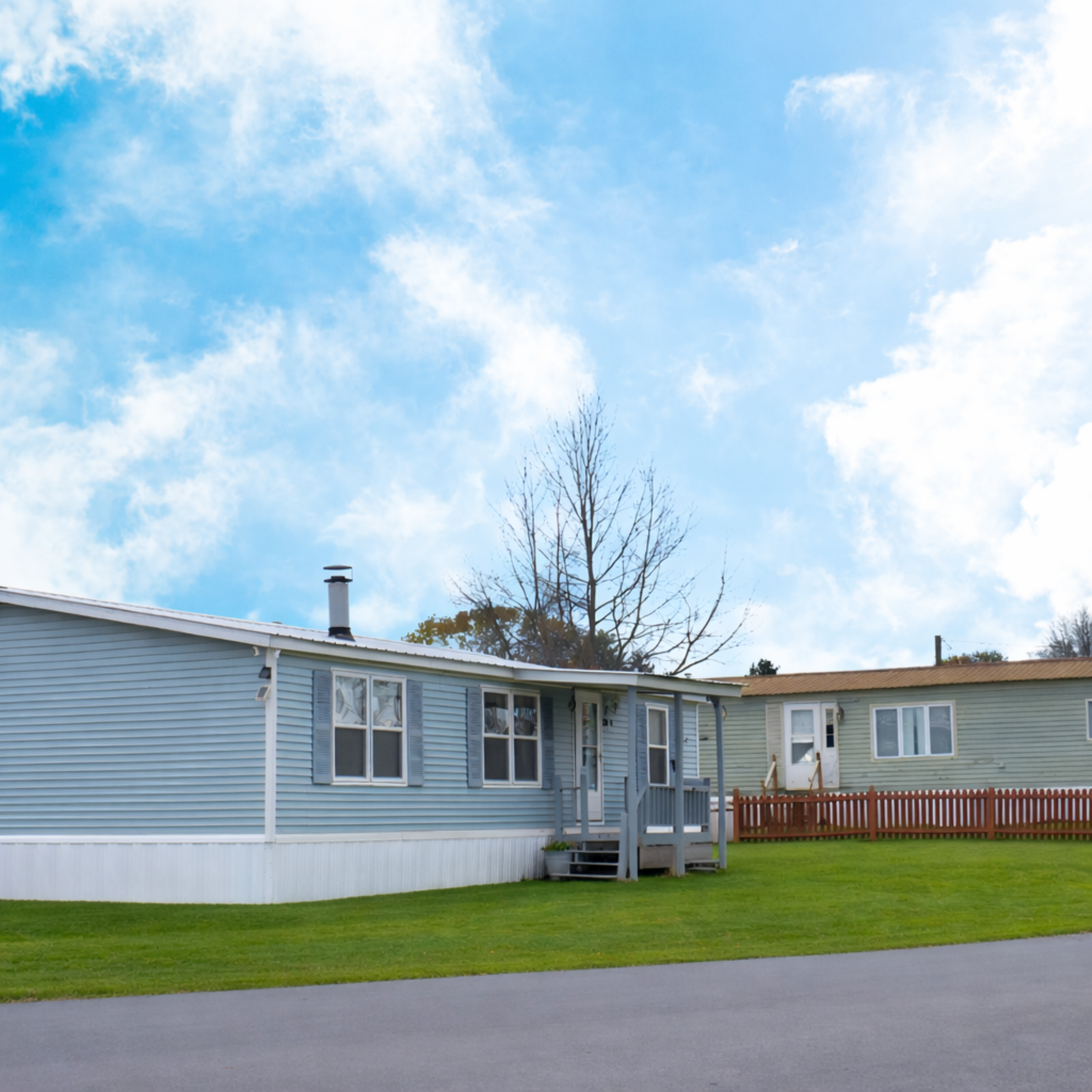 A row of light blue and green mobile homes with small front steps, surrounded by green grass, under a blue sky with scattered clouds.