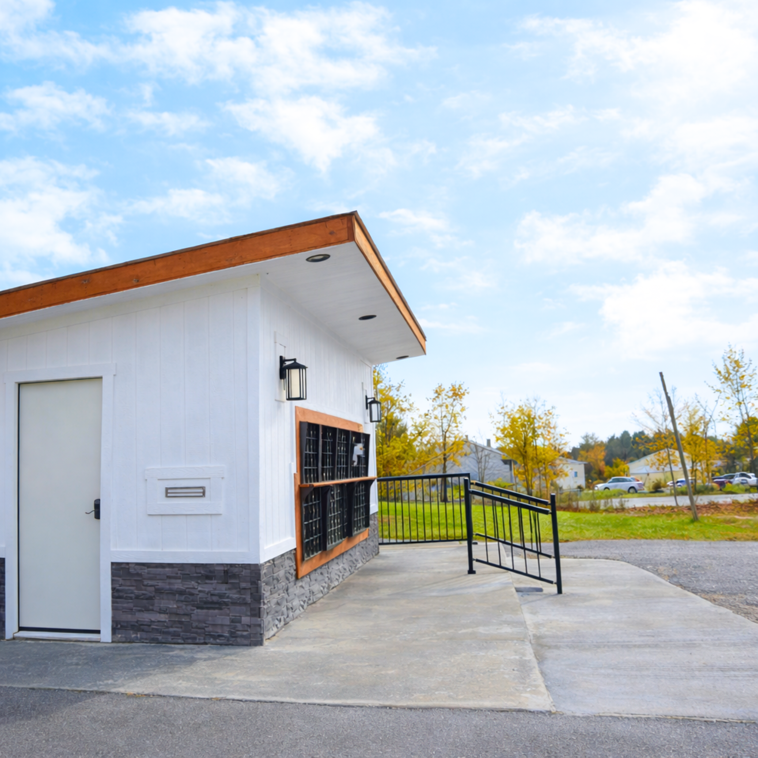 A small, modern building with white exterior walls, a stone base, and a flat roof with wood trim. There is a door and a window with black metal bars, and outdoor wall-mounted lights. The building is situated near a sidewalk, with trees and parked car