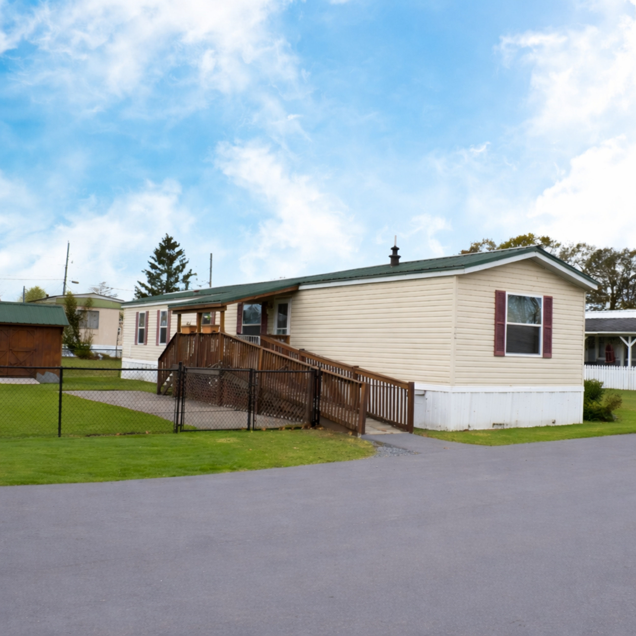 Single-story beige mobile home with purple shutters, brown ramp, chain link fence, green lawn, and blue sky with clouds.